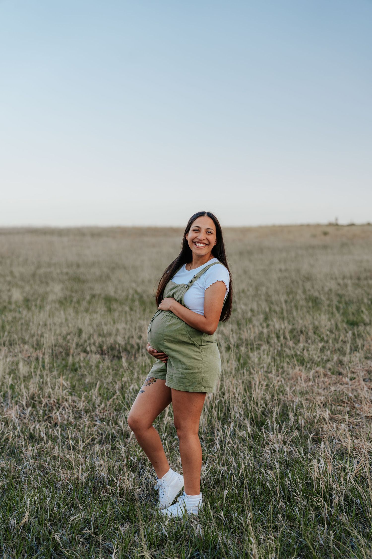 Smiling pregnant woman in green overalls holding baby bump in an open field.