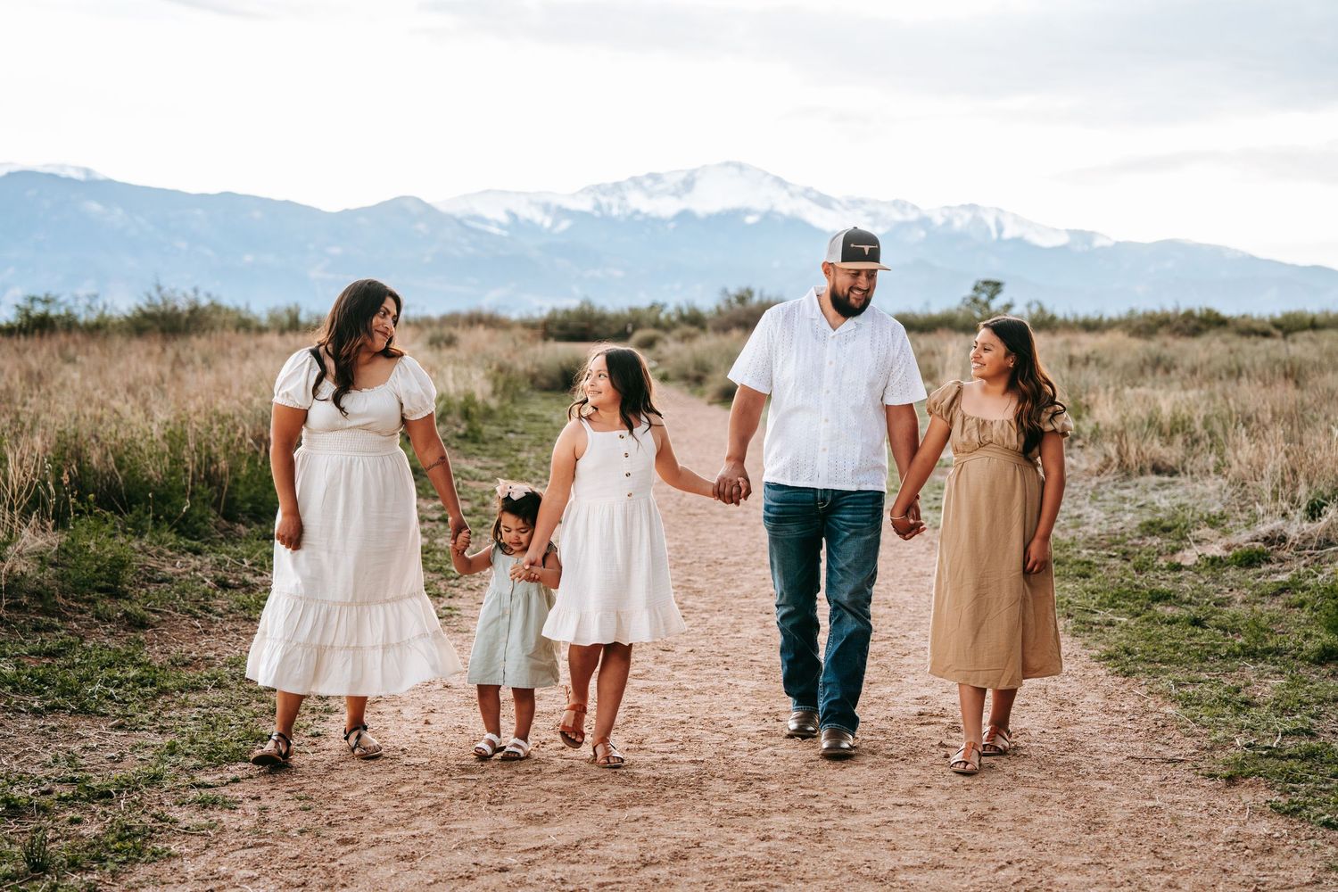 Family of five walking hand-in-hand on dirt path with snow-capped mountains in the background.