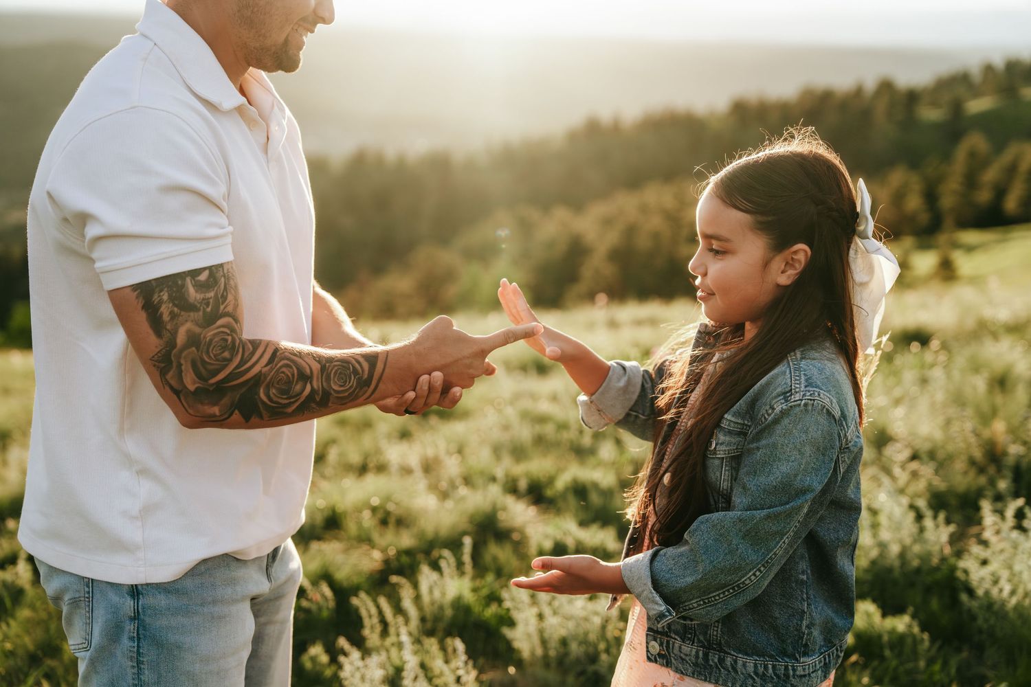 Tattooed father showing something small to his daughter in a sunlit green meadow.