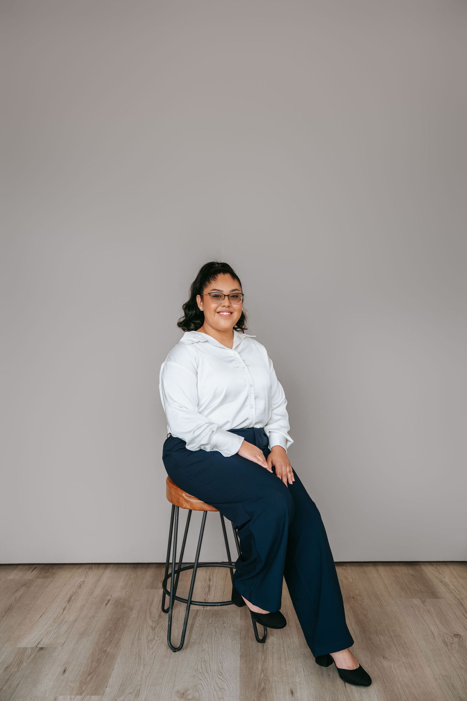 Woman in white shirt and navy pants sitting on a stool wearing glasses against a gray background.