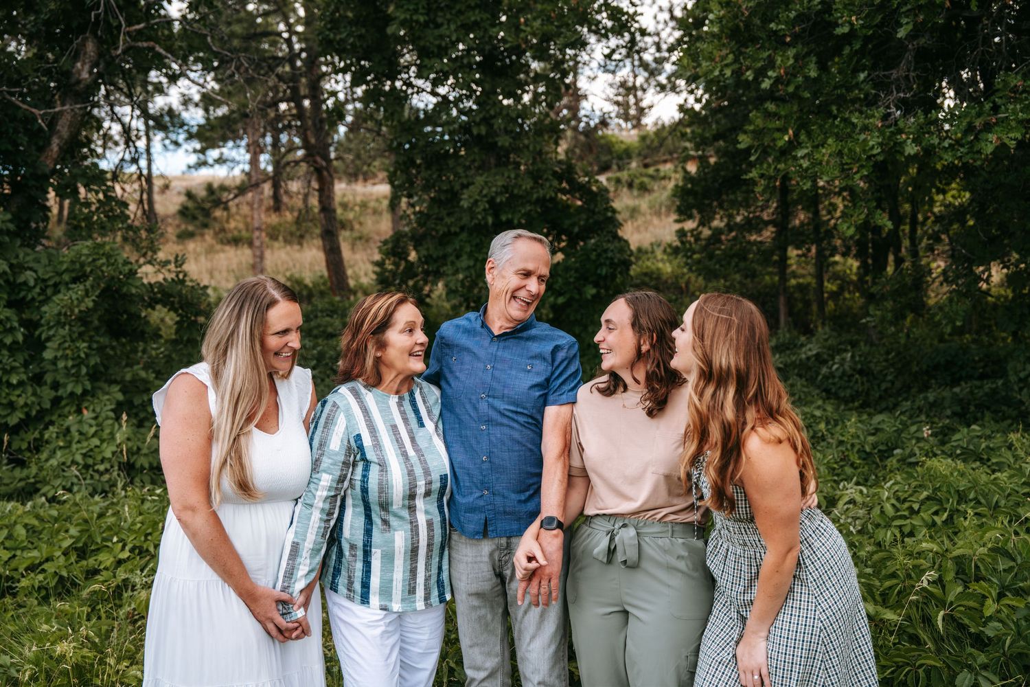 A happy family of five smiling and laughing together outdoors surrounded by lush green trees.