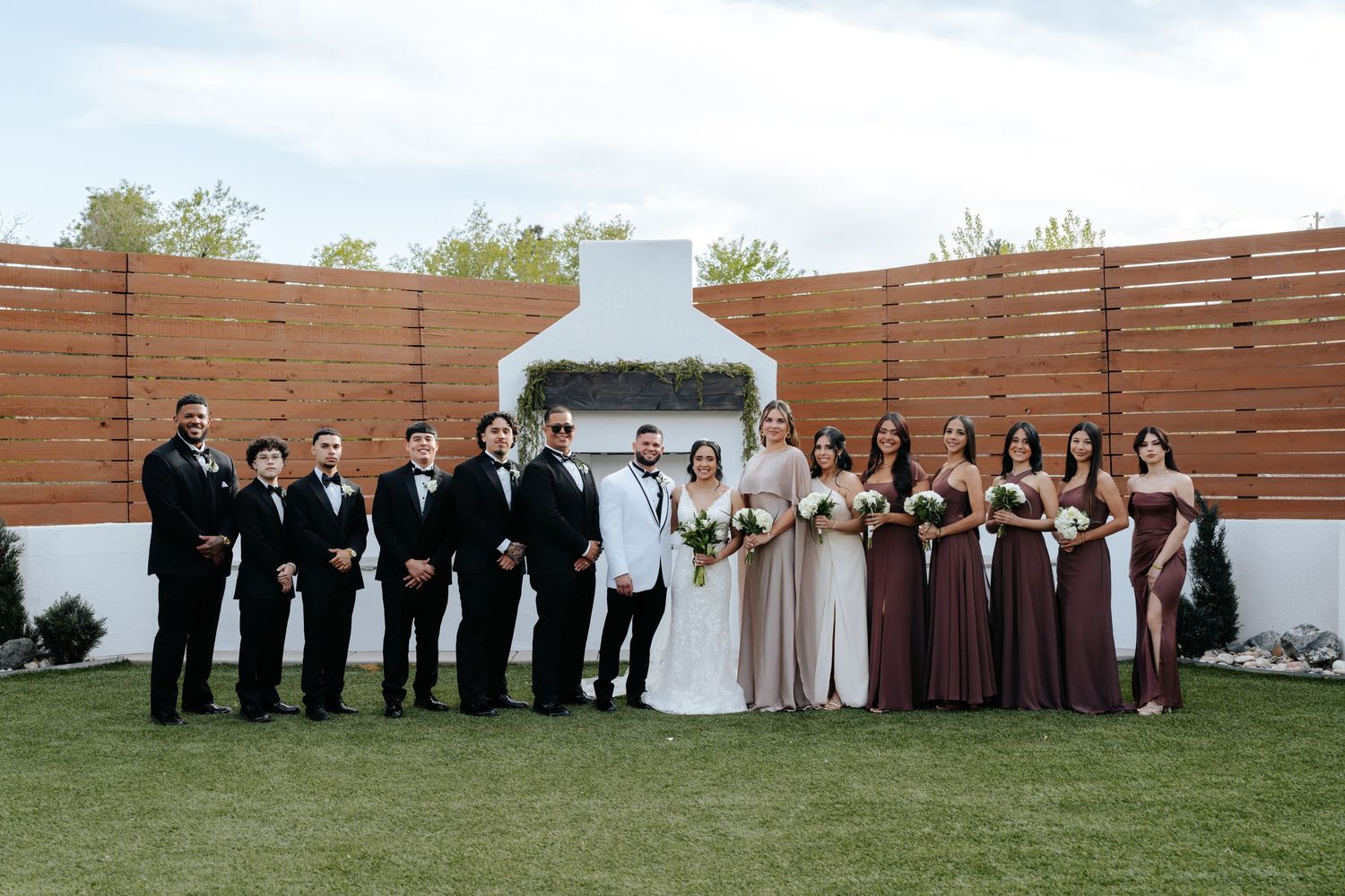 Wedding party poses outdoors with groomsmen in black tuxedos and bridesmaids in mauve gowns before a white fireplace backdrop.