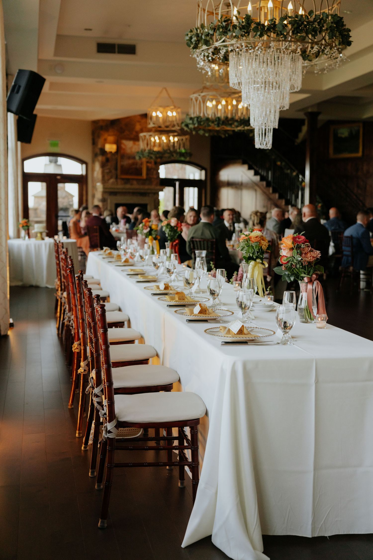 Elegant wedding reception hall with long white tables, chiavari chairs, and crystal chandeliers adorned with greenery.