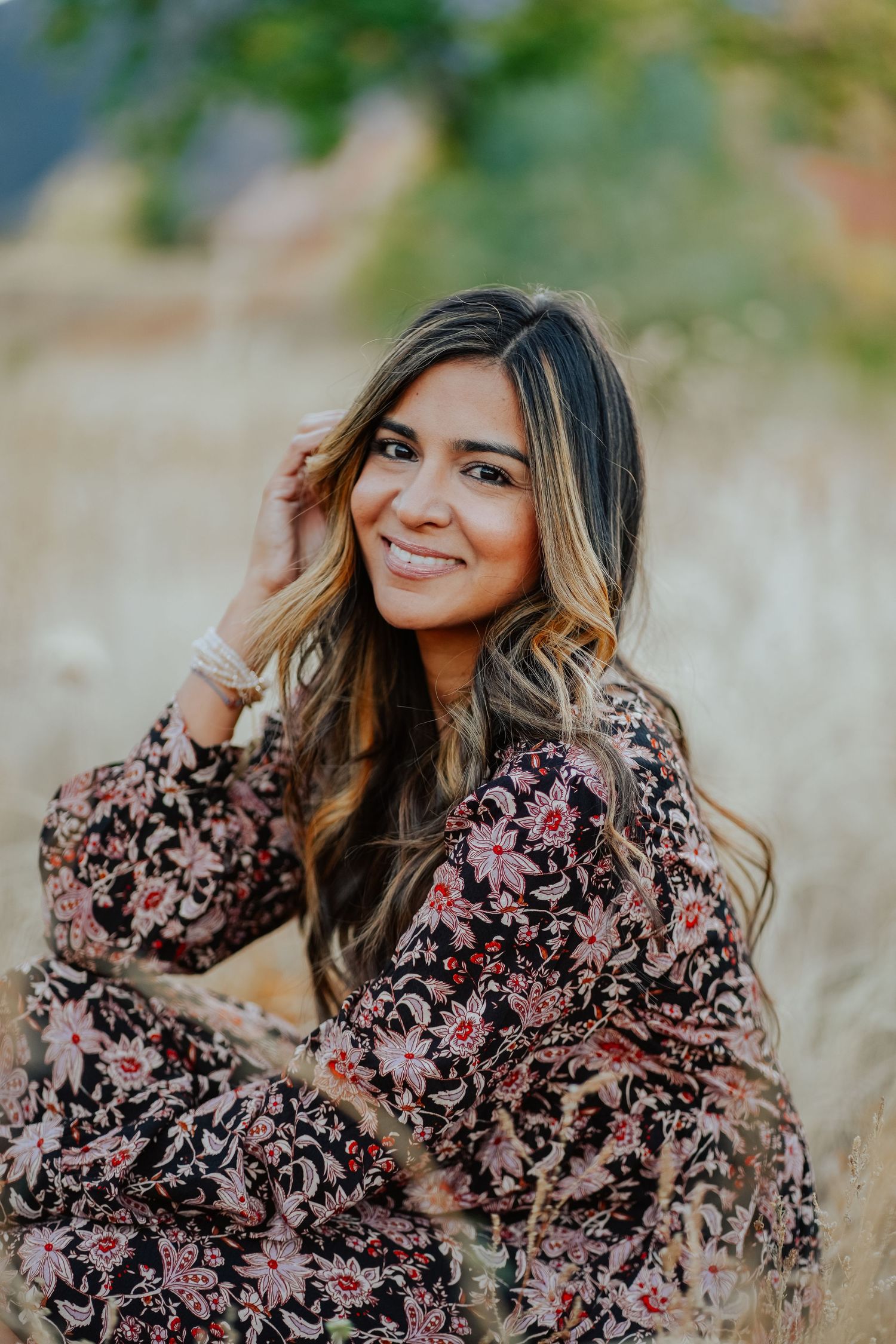 Woman in black floral dress sitting in dry grass field touching hair and smiling.