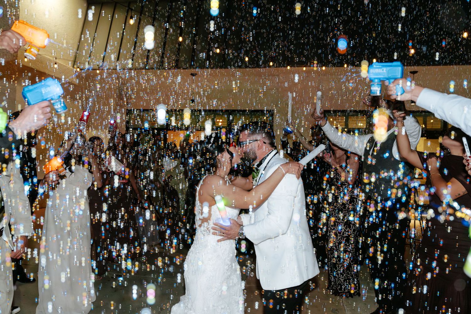 Newlyweds share romantic first dance surrounded by falling bubbles and cheering guests at wedding reception celebration.