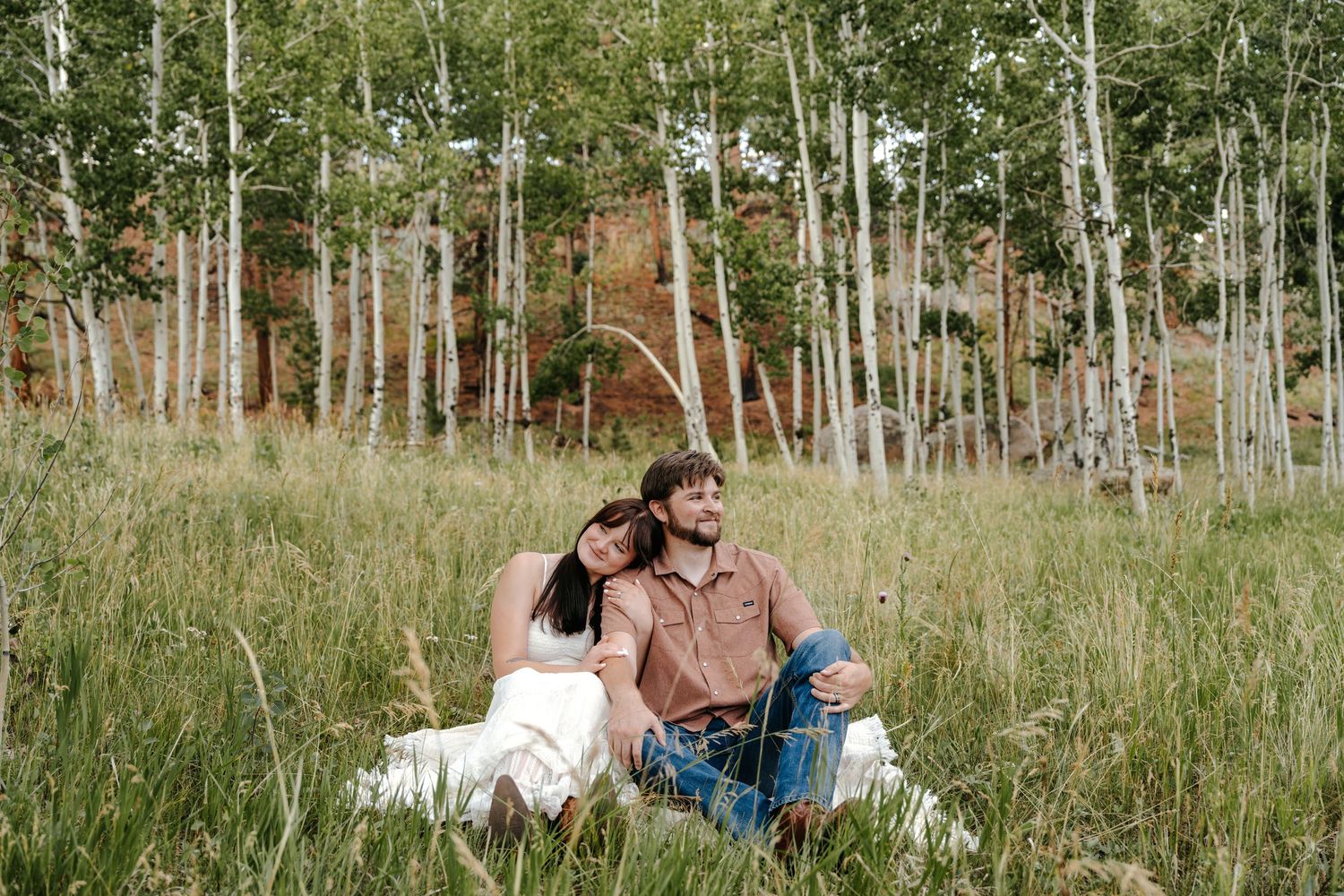 Couple sitting together in a lush meadow surrounded by aspen trees and red rock cliffs.