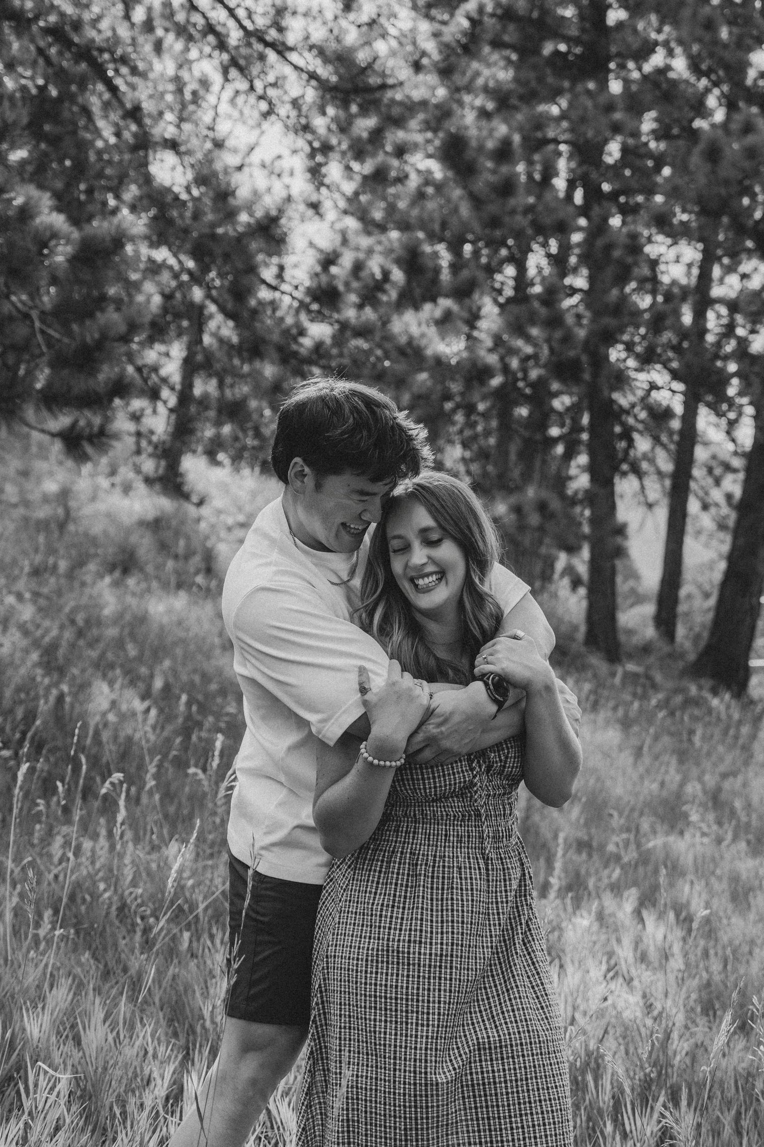 Black and white photo of a happy couple embracing and laughing together in a lush forest meadow.