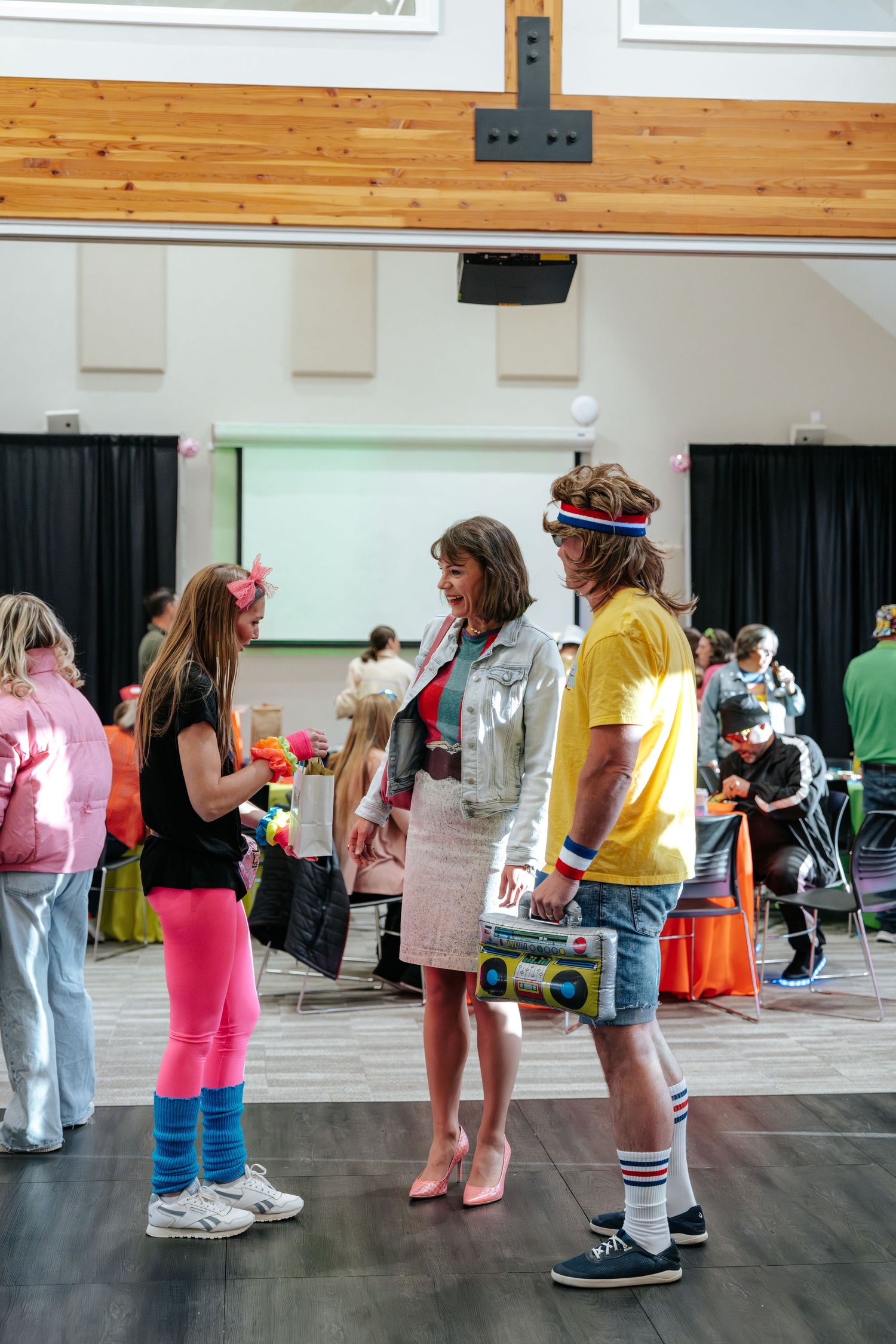 People in colorful 80s costumes chat at an indoor event, featuring bright leg warmers, headbands, and a boombox prop.