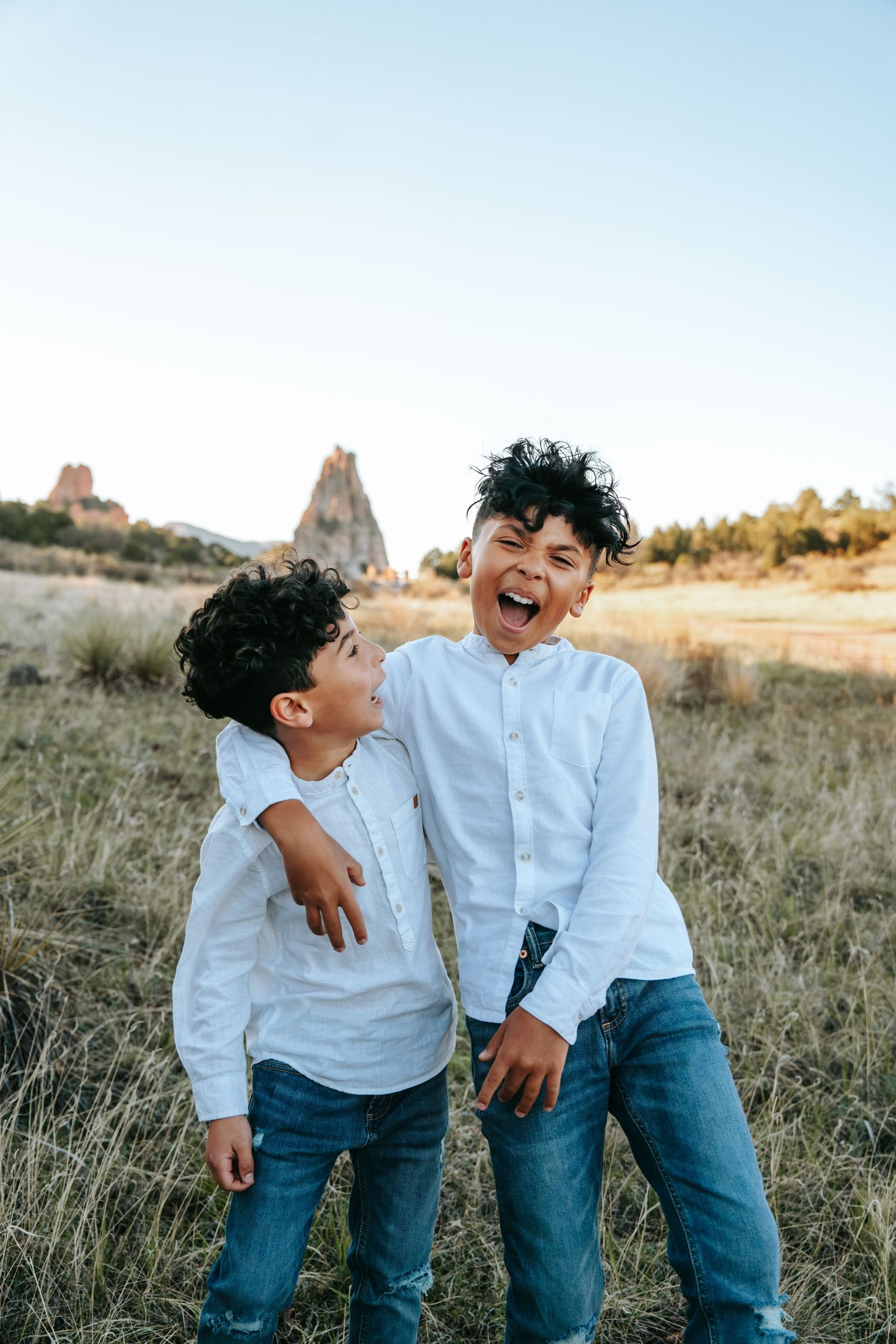 Two laughing boys in matching white shirts and jeans playfully embracing in a field with rocky landscape.