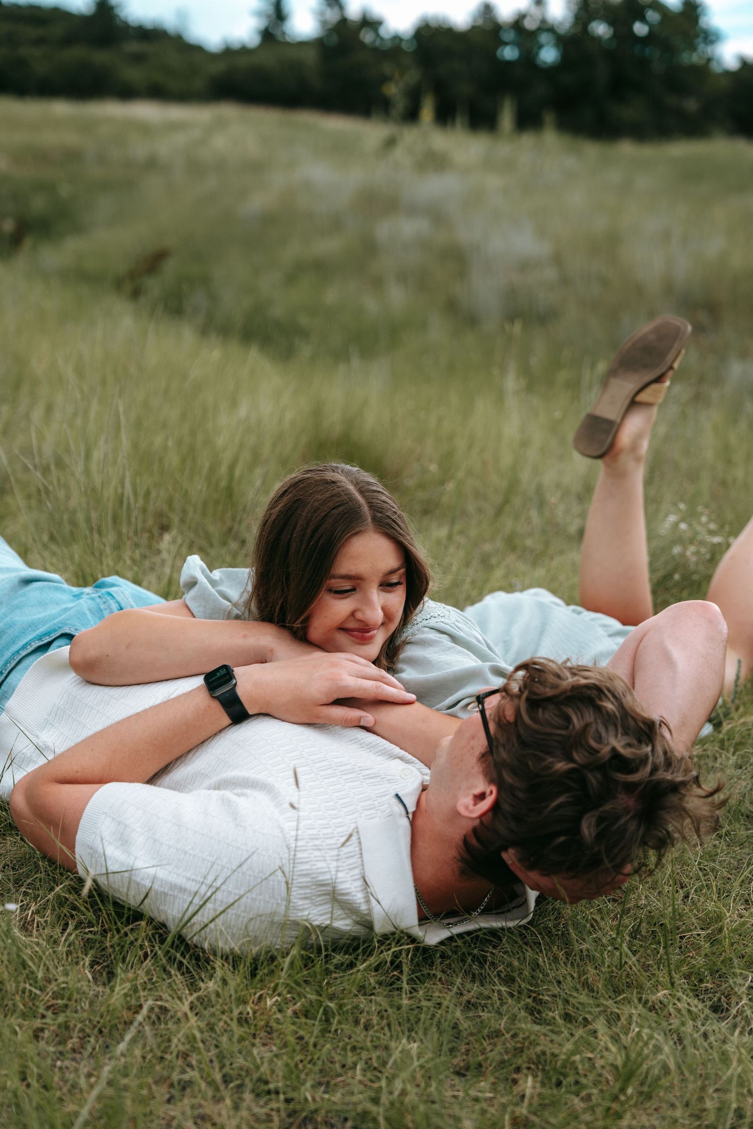 Couple lying together in lush green grass, laughing and enjoying a playful outdoor moment.