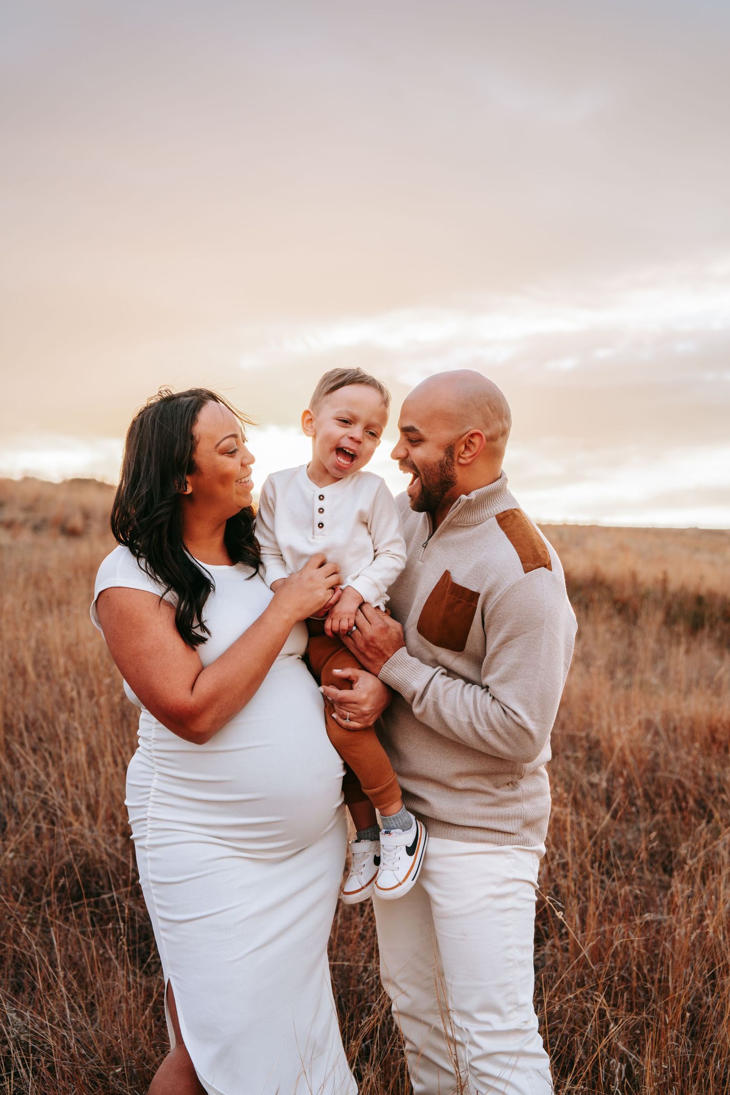 Happy family maternity photo in golden field at sunset with pregnant mother in white dress and toddler son.