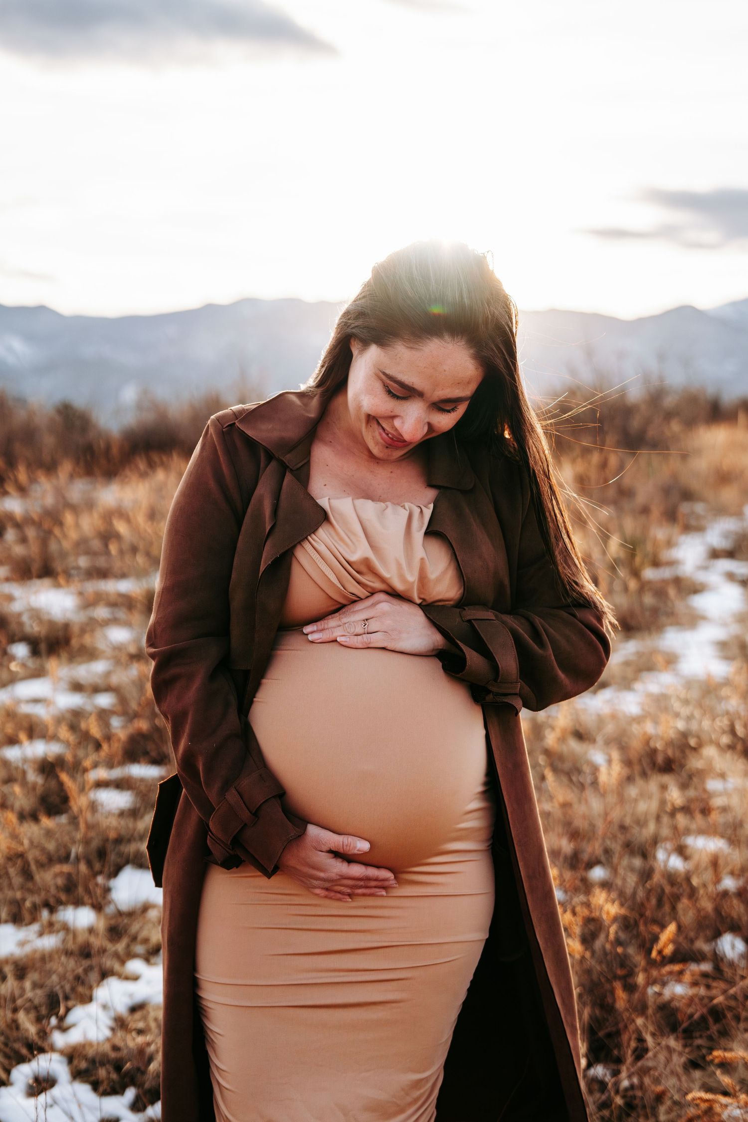 Glowing pregnant woman in beige dress and brown coat smiling down at baby bump in sunny outdoor field.