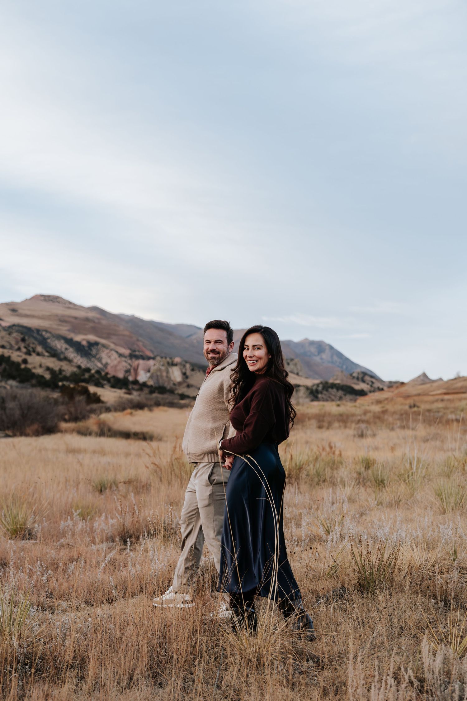 Couple posing together in golden autumn field with mountain backdrop during fall engagement photo session.