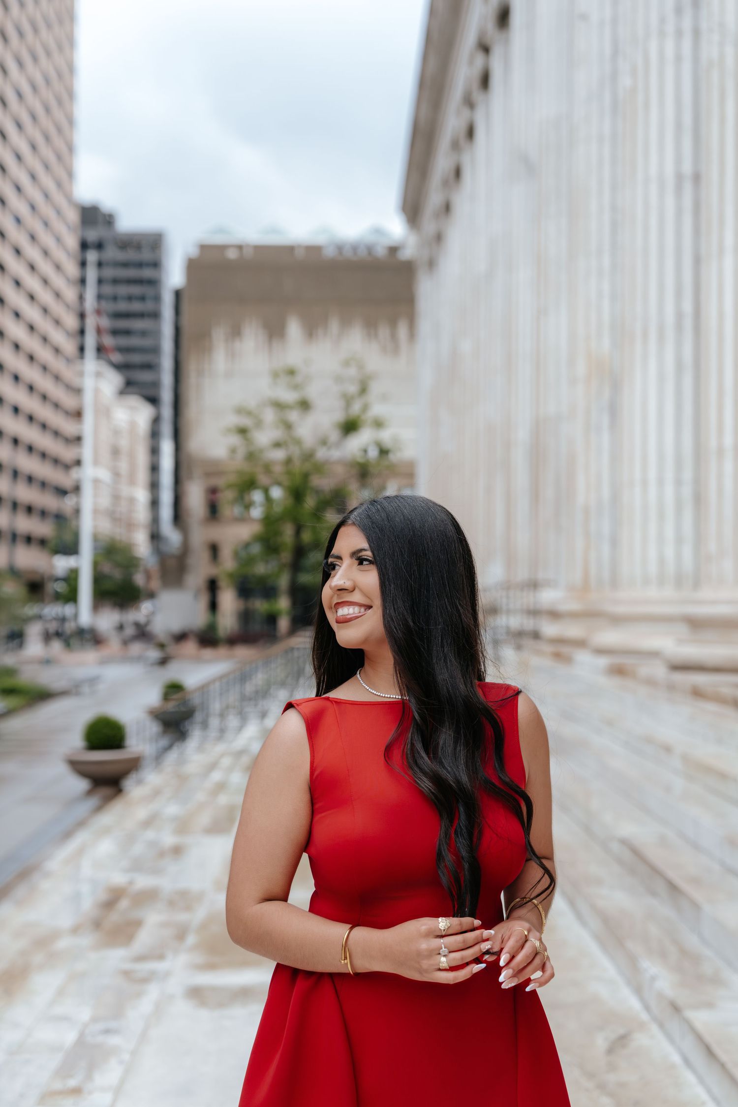 Young woman in red dress smiling outdoors near classical building columns in urban setting.
