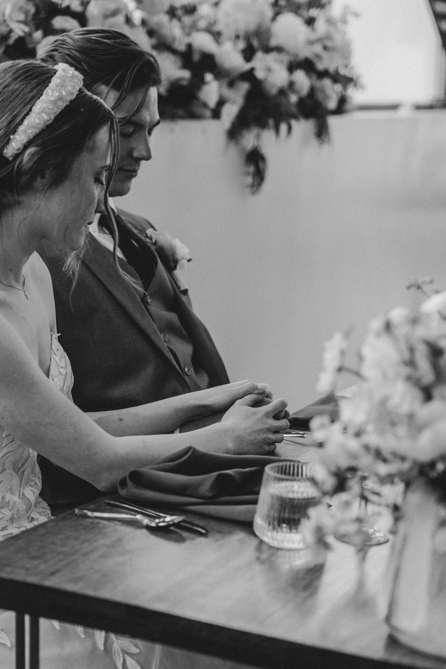 Black and white photo of a bride and groom holding hands at their sweetheart table with floral arrangements.
