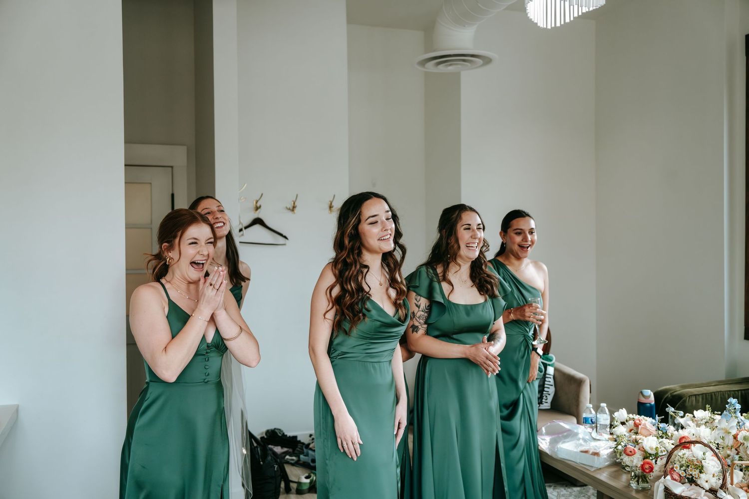 Four bridesmaids in elegant green satin dresses reacting with joy and excitement during wedding preparations.