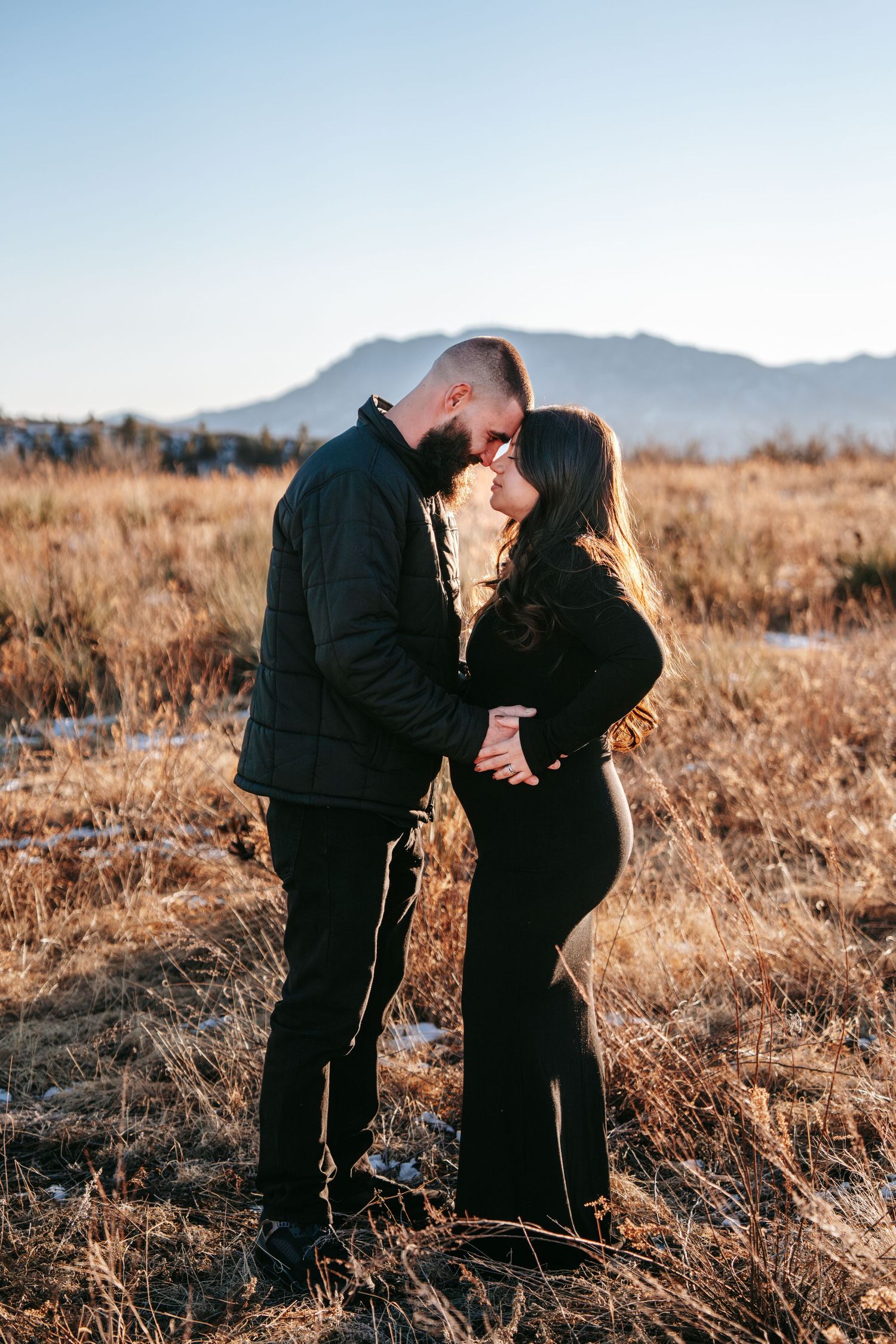 Couple nearly kissing during golden hour maternity session in dry grass field with mountain views.