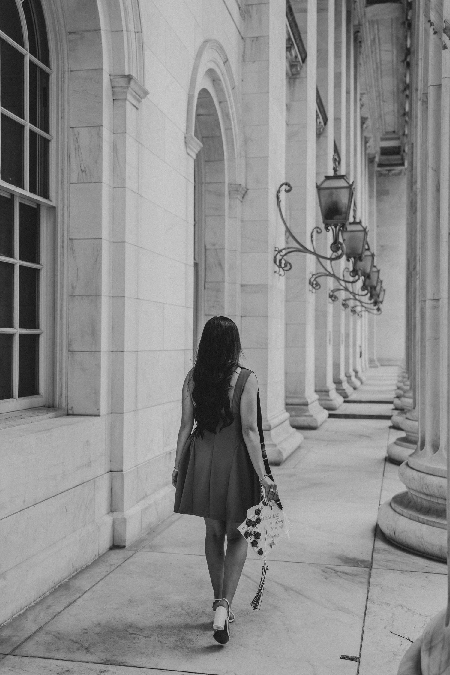 Black and white photo of a young woman walking away down a columned corridor holding a bouquet of flowers.