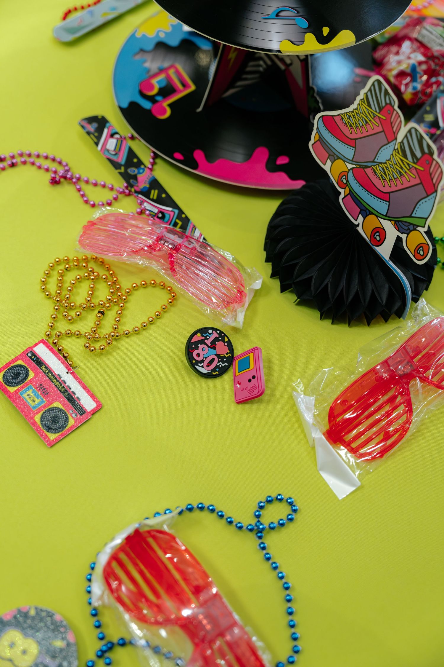 Colorful 80s retro party accessories on yellow background, including bead necklaces, sunglasses, pins, and vinyl record.