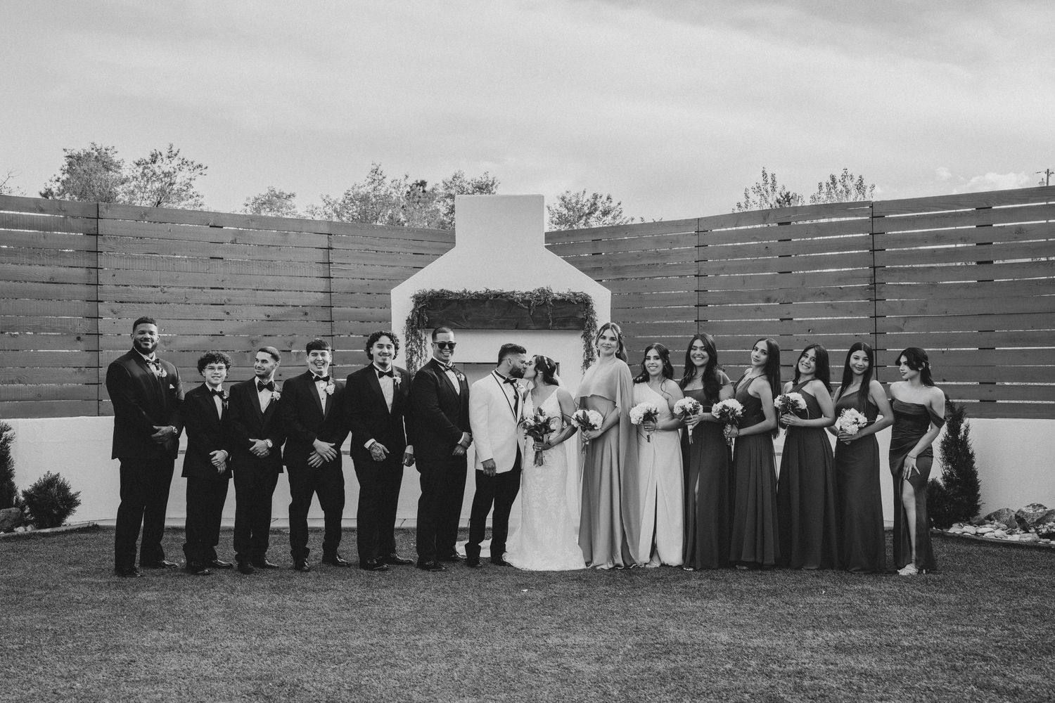 Black and white wedding party photo with bride, groom, groomsmen, and bridesmaids posed in front of outdoor fireplace.