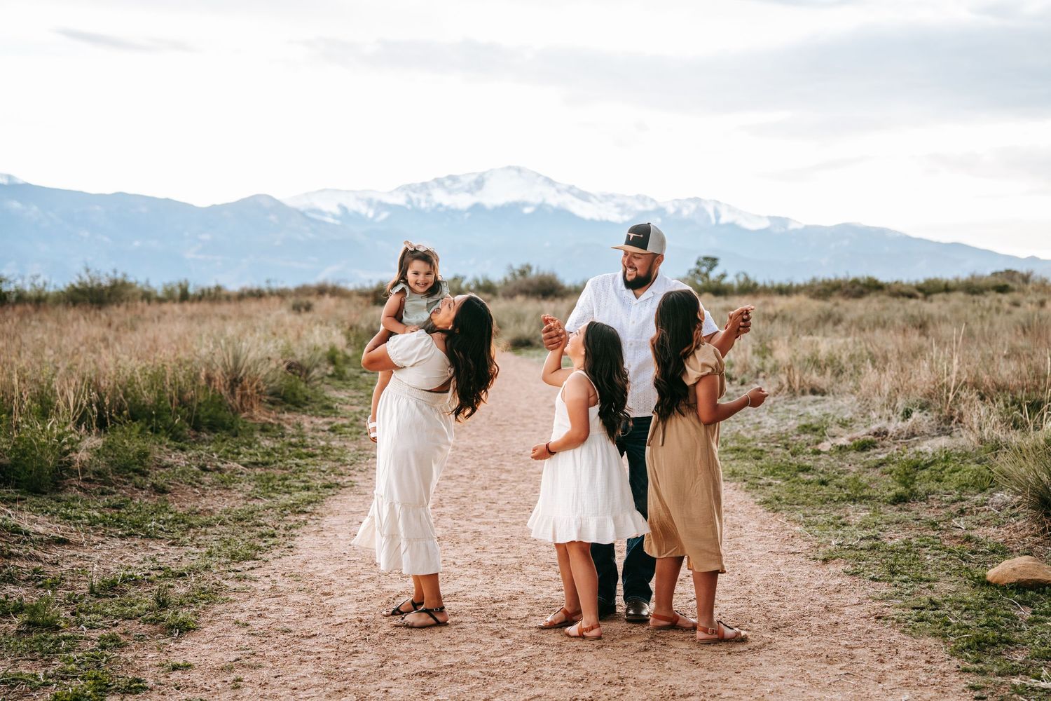 Family of five standing together on a dirt path surrounded by tall grass and mountain scenery.
