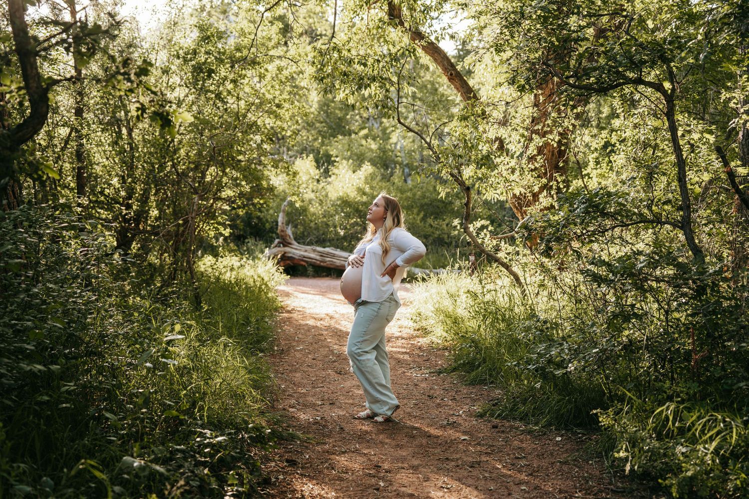 Woman in white outfit walking on a sunny forest trail surrounded by lush green trees.