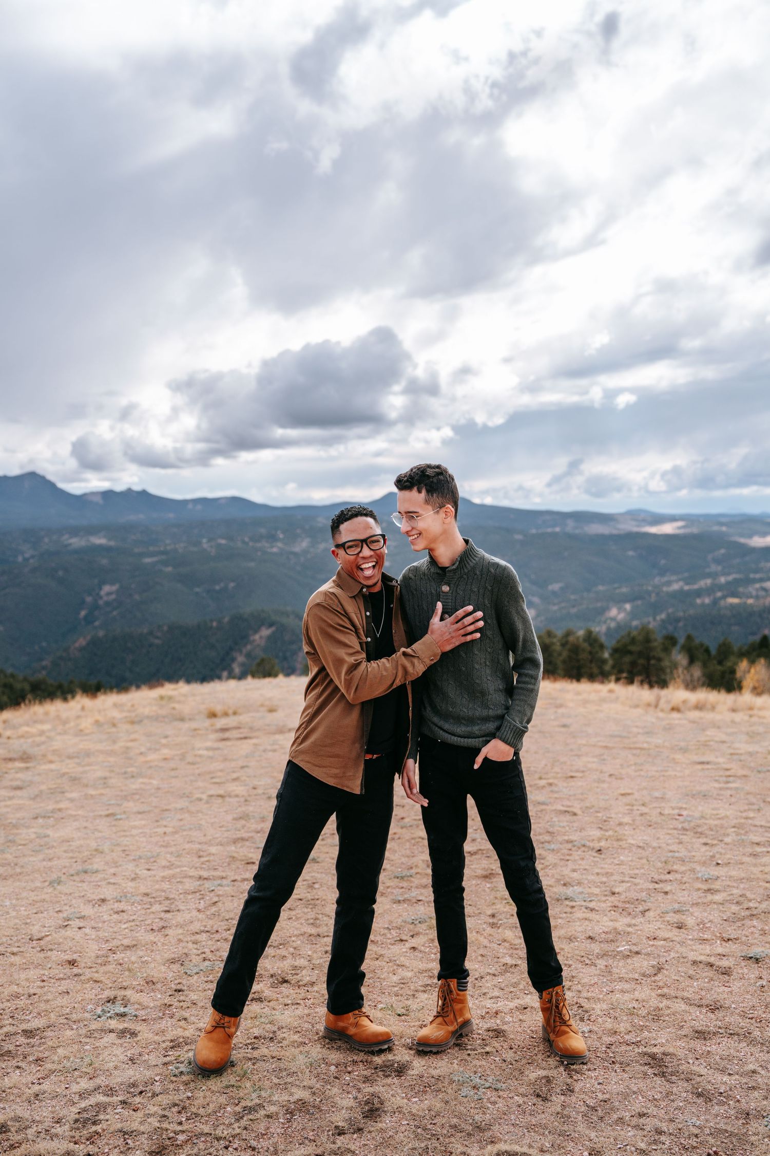 Two young men laughing and embracing on a hilltop with dramatic mountain and cloudy sky backdrop.