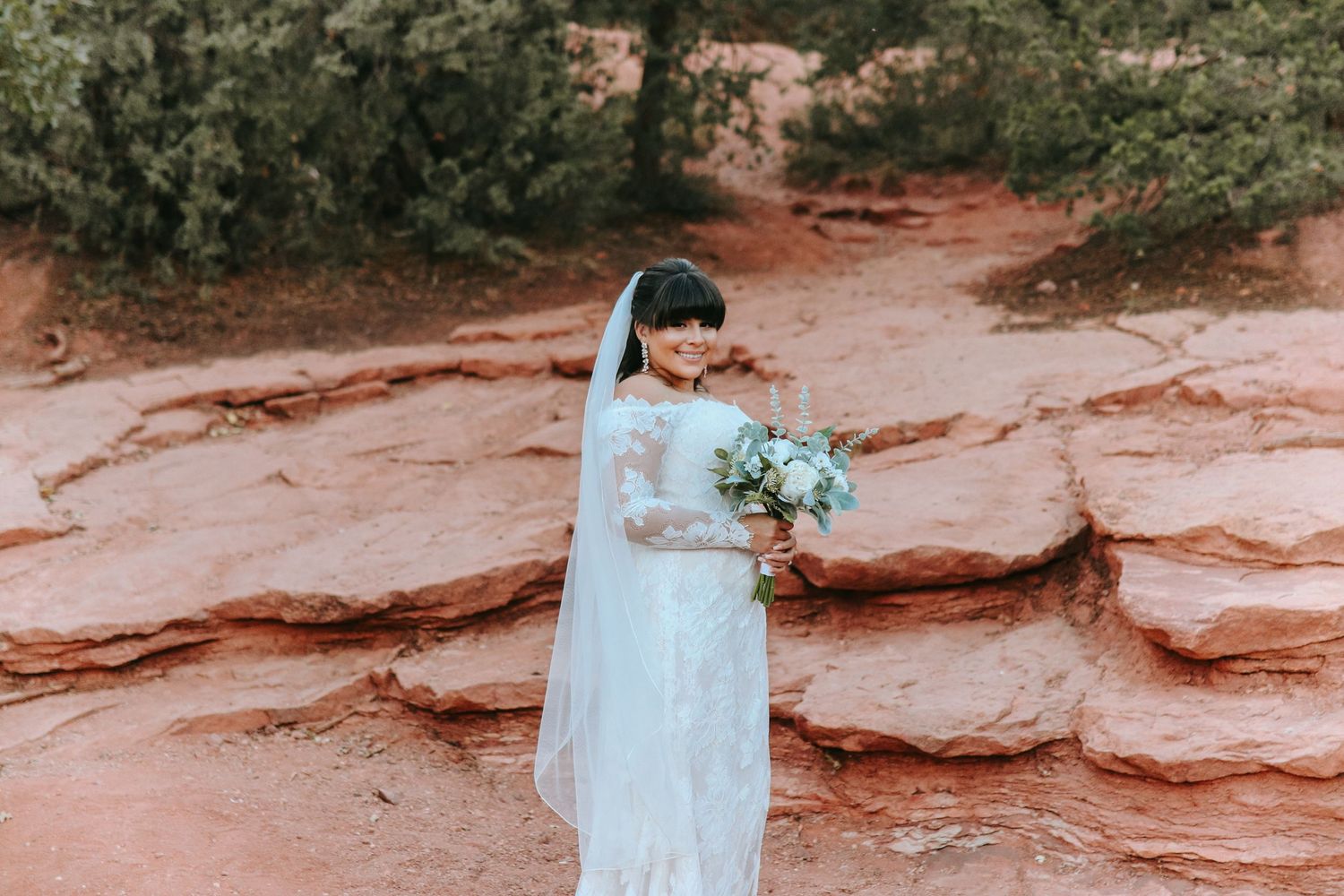 Bride in lace wedding gown holding white bouquet standing on red rocks outdoors.