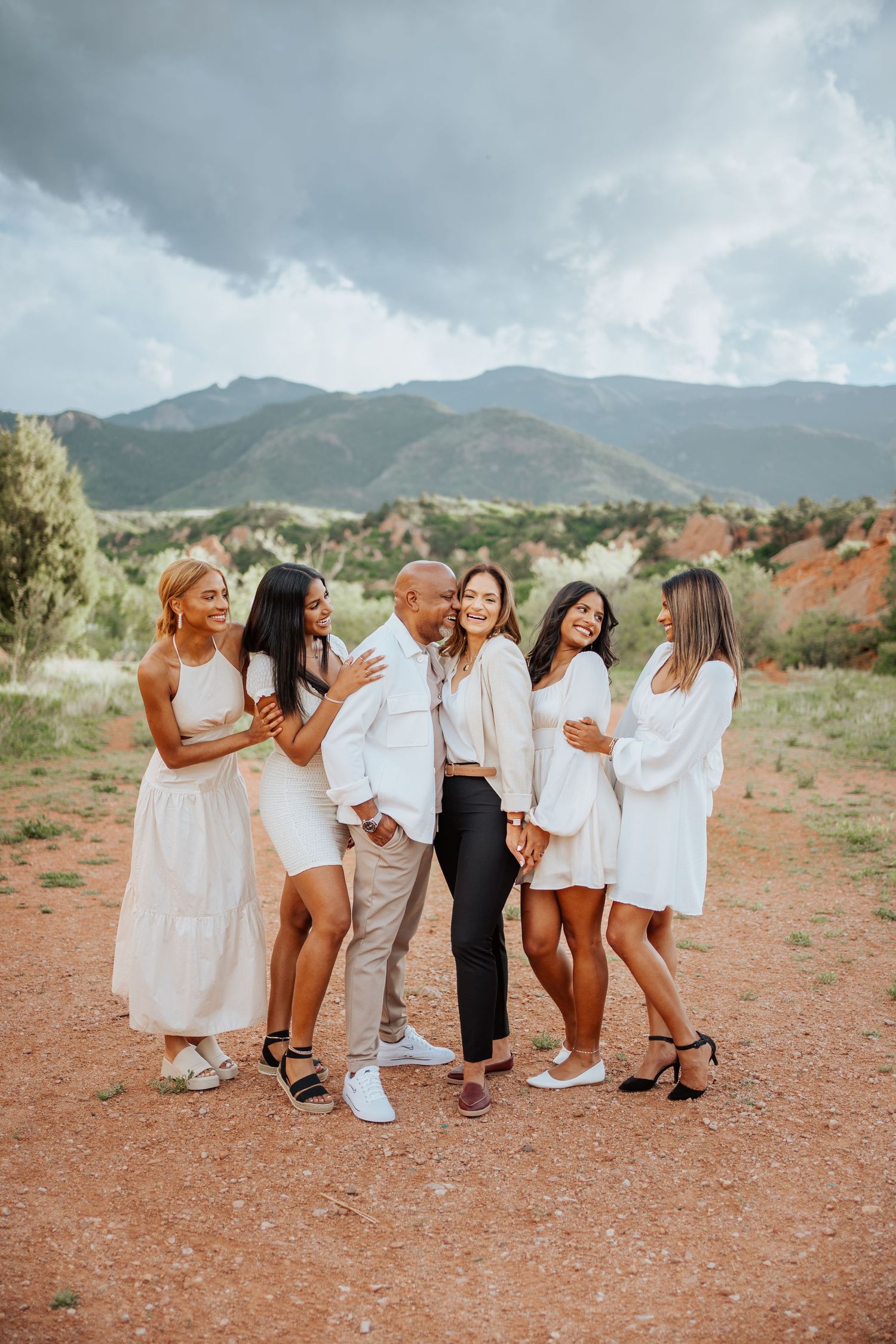 Smiling family group hugging in white outfits on red dirt trail with mountain views.