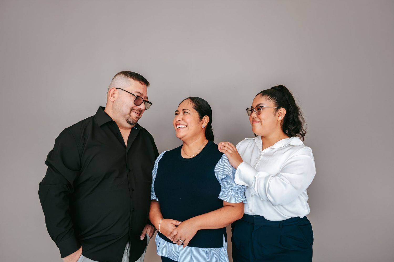 Three smiling people laughing together in a casual group portrait against a gray background.