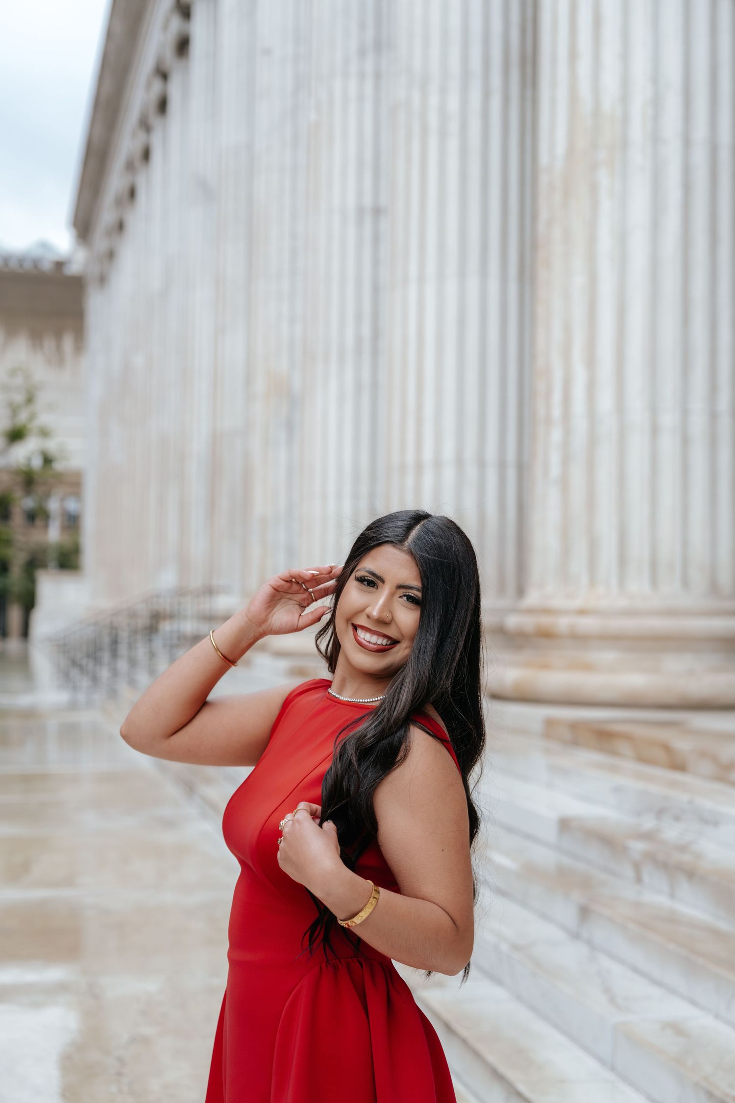Smiling young woman in red dress poses gracefully beside tall marble columns, touching her hair with a gold bracelet.