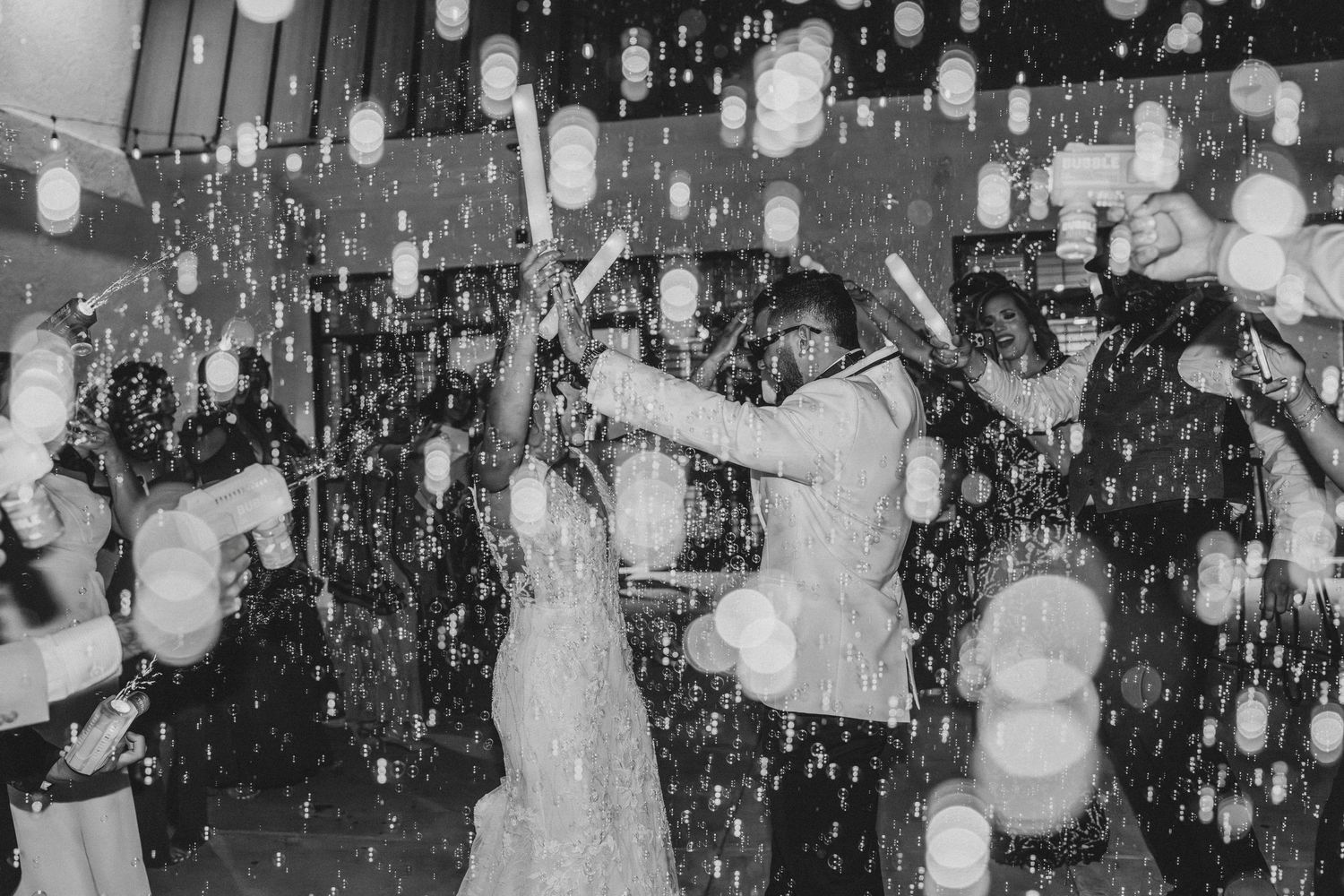 Black and white wedding photo of couple dancing joyfully amid falling confetti and glowing candles held by guests.