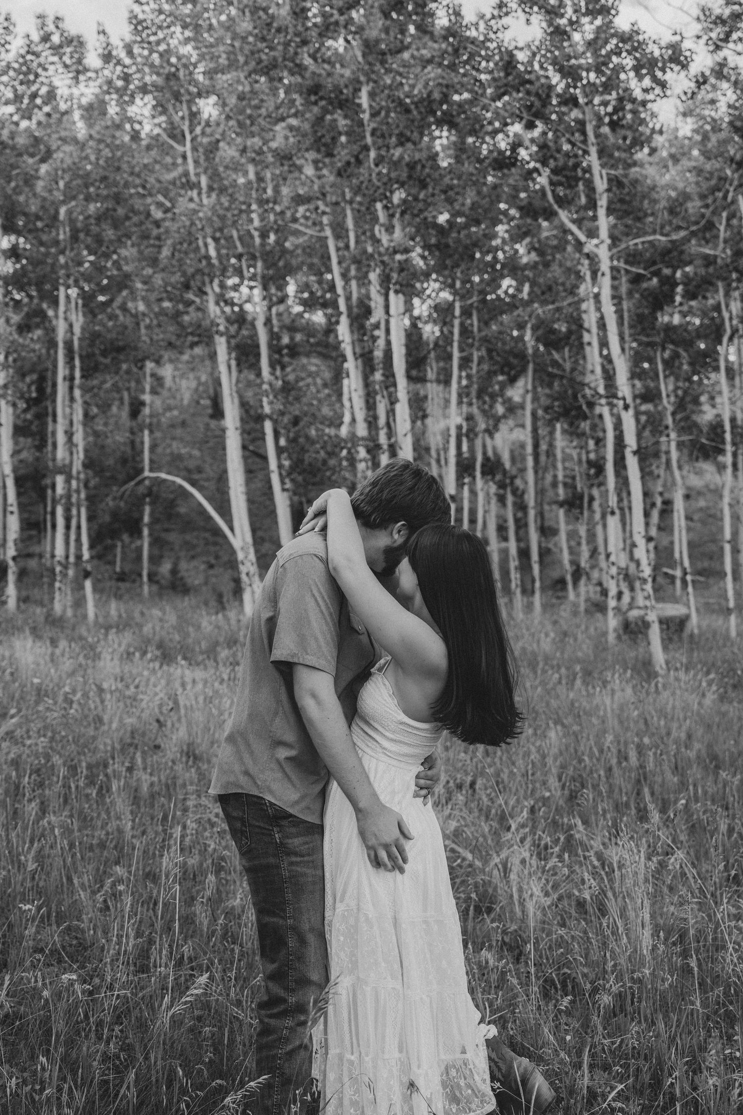 Black and white photo of a couple kissing in a meadow surrounded by birch trees.