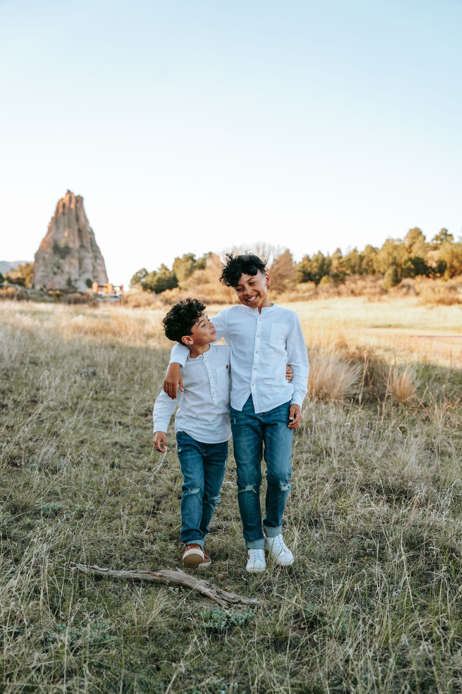 Two young brothers in white shirts hugging and smiling at each other in an open field with a rock formation.