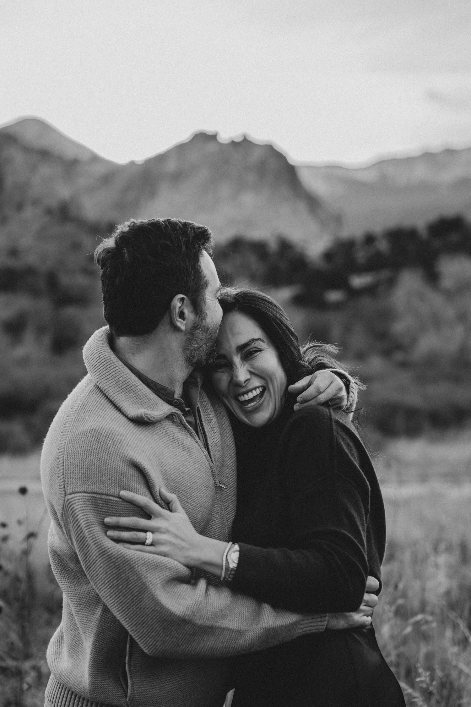 Black and white photo of a laughing couple embracing in a mountain meadow during an outdoor engagement session.