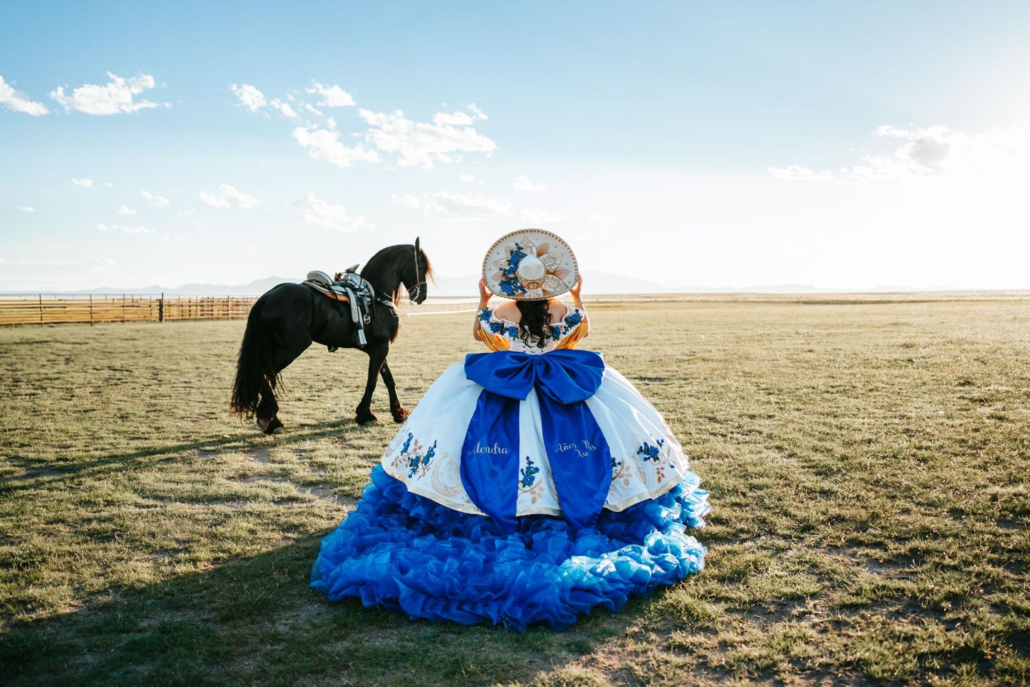Quinceañera in blue white ball gown with sombrero poses near black horse in open field.