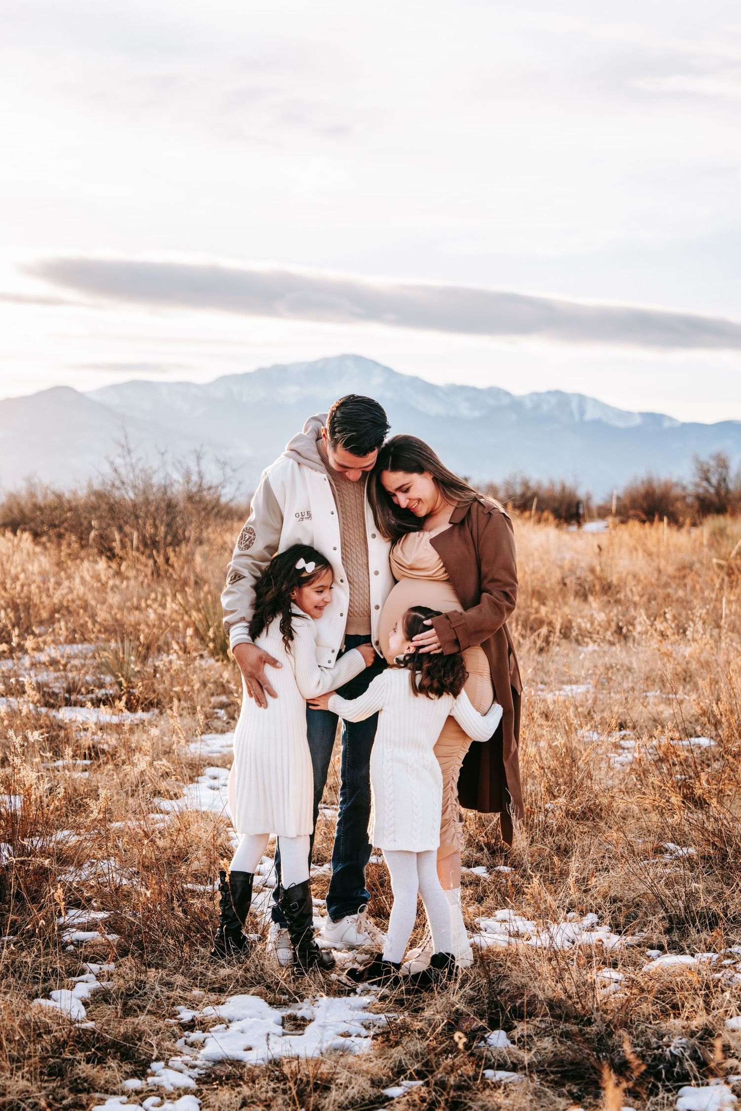 Family of four gathered together in snowy field, parents and two daughters admiring pregnant mothers baby bump.