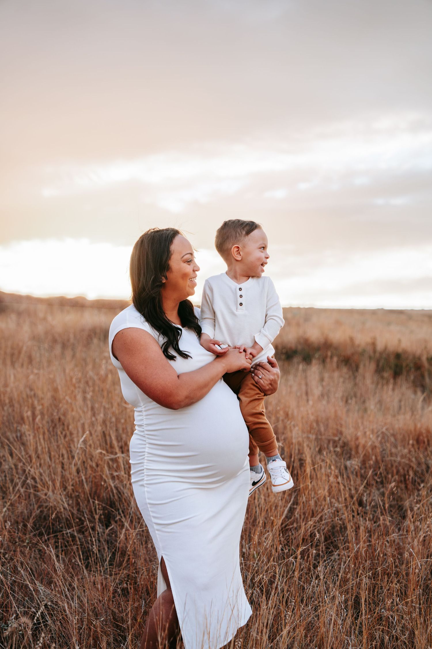 Pregnant woman in white dress holds her toddler son in a golden grass field at sunset.