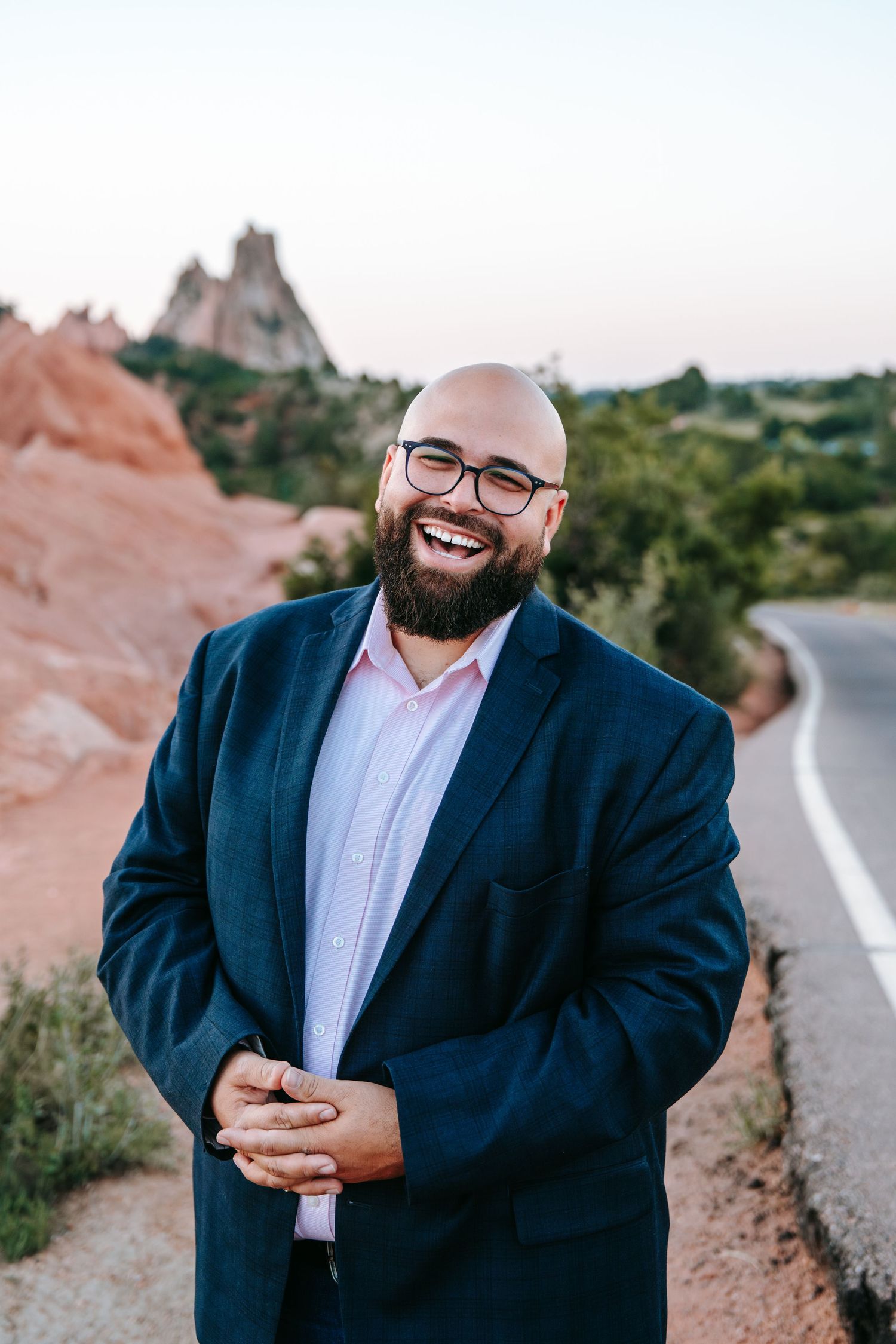 Laughing bald bearded man in navy blazer posing along scenic red rock road at dusk.