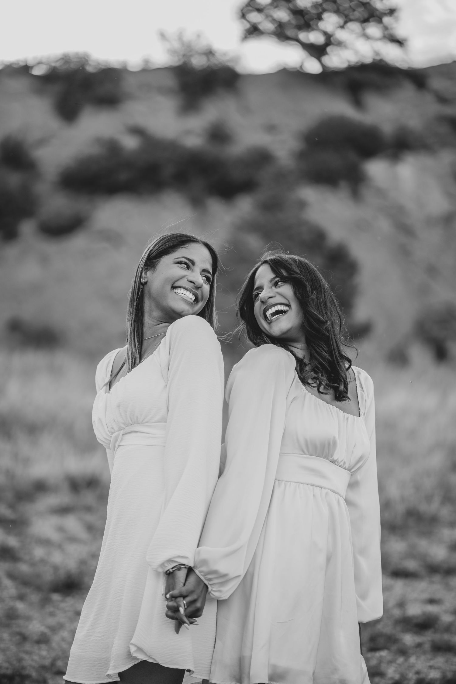 Black and white portrait of two smiling women in white dresses holding hands outdoors.