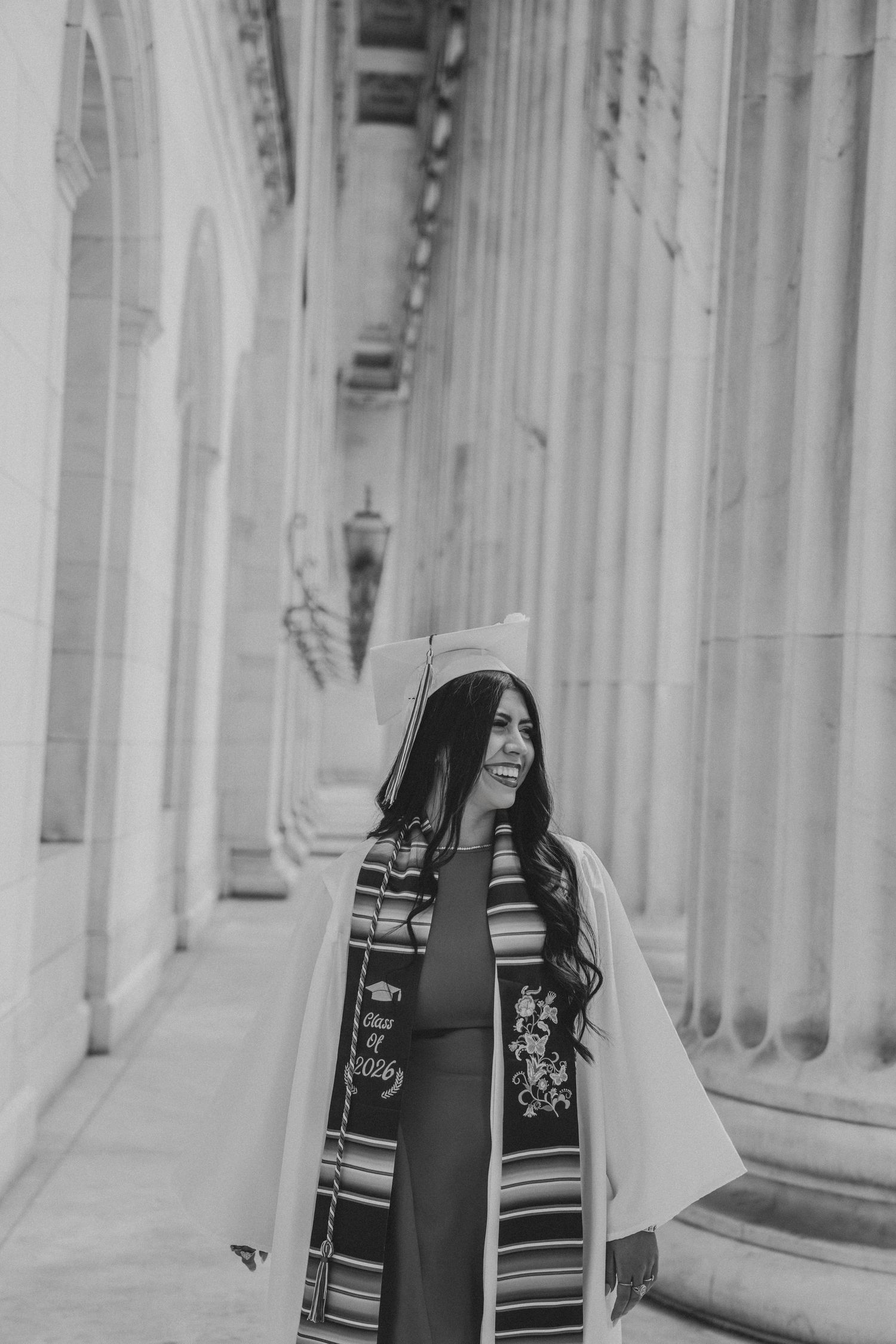 Black and white photo of a graduate in cap and gown with serape stole walking between large stone columns.
