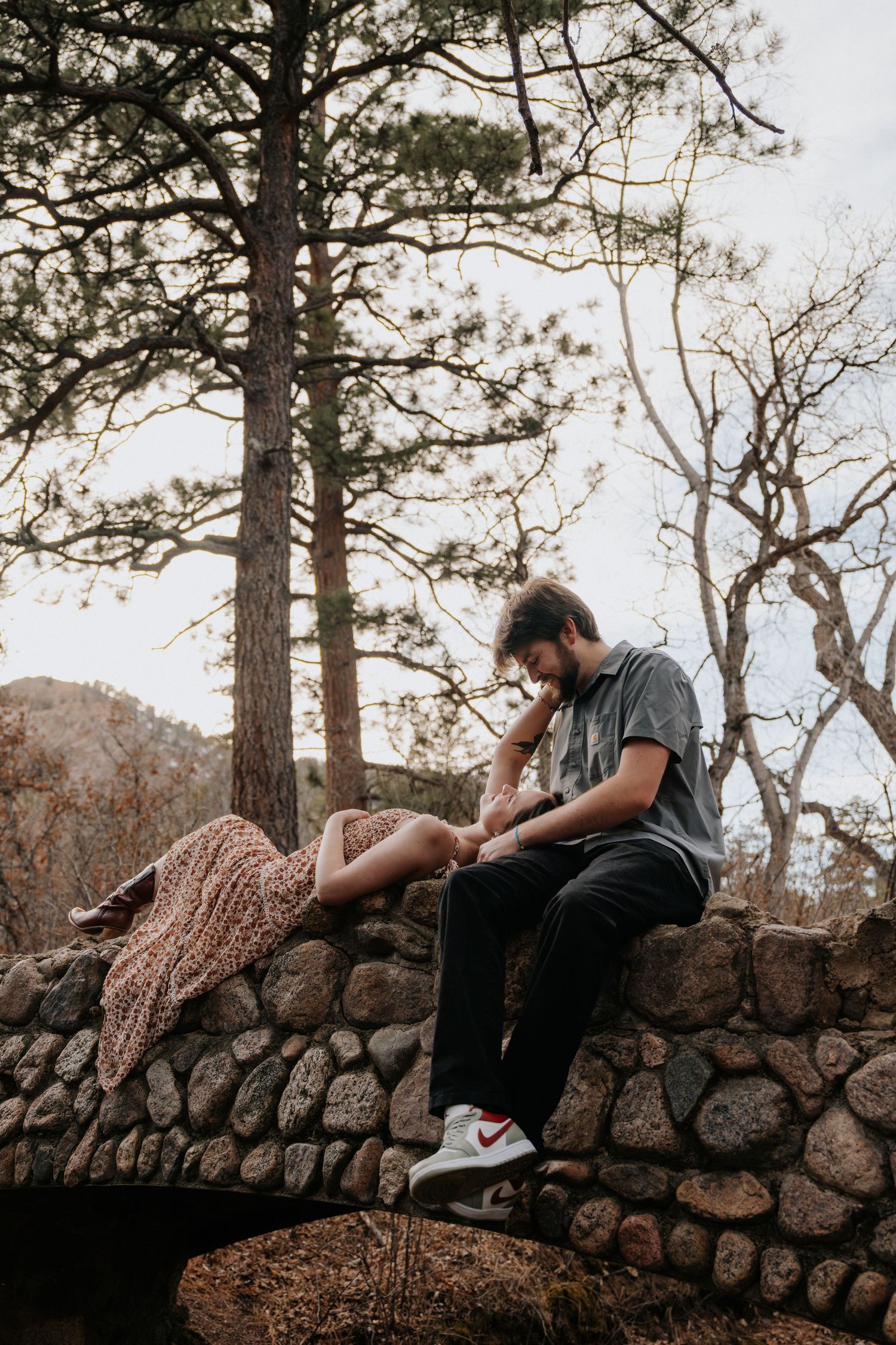 Couple sitting on rustic stone wall in forest, man tenderly holding womans hand outdoors.