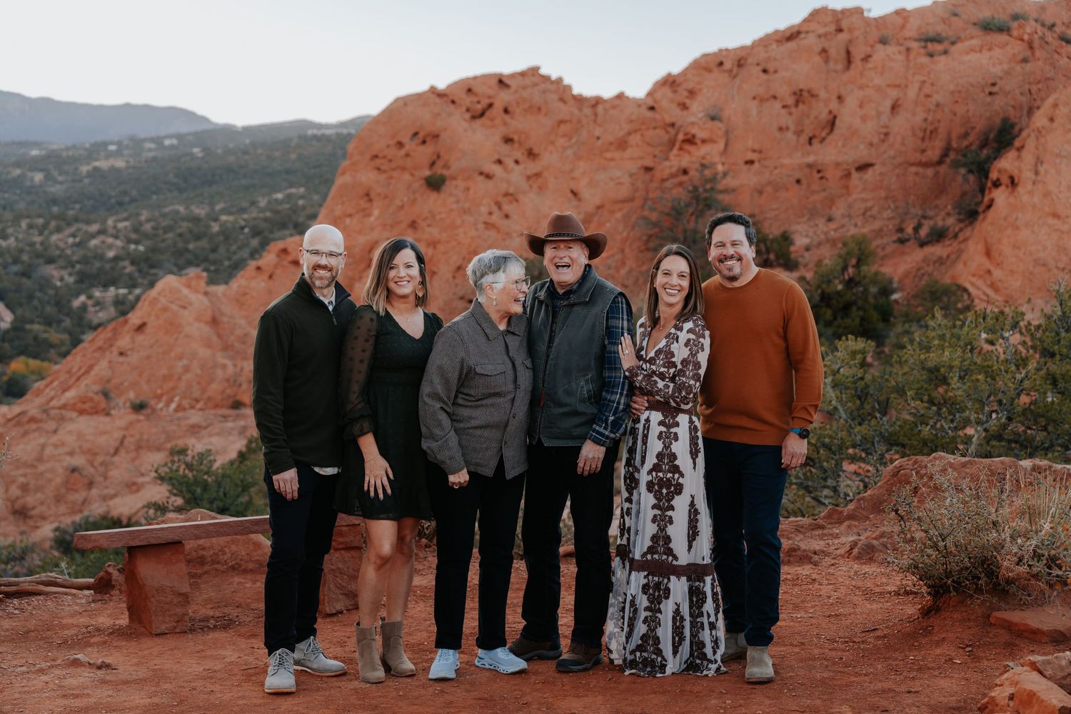 Six family members smiling together outdoors with towering red rock formations and blue sky in the background.