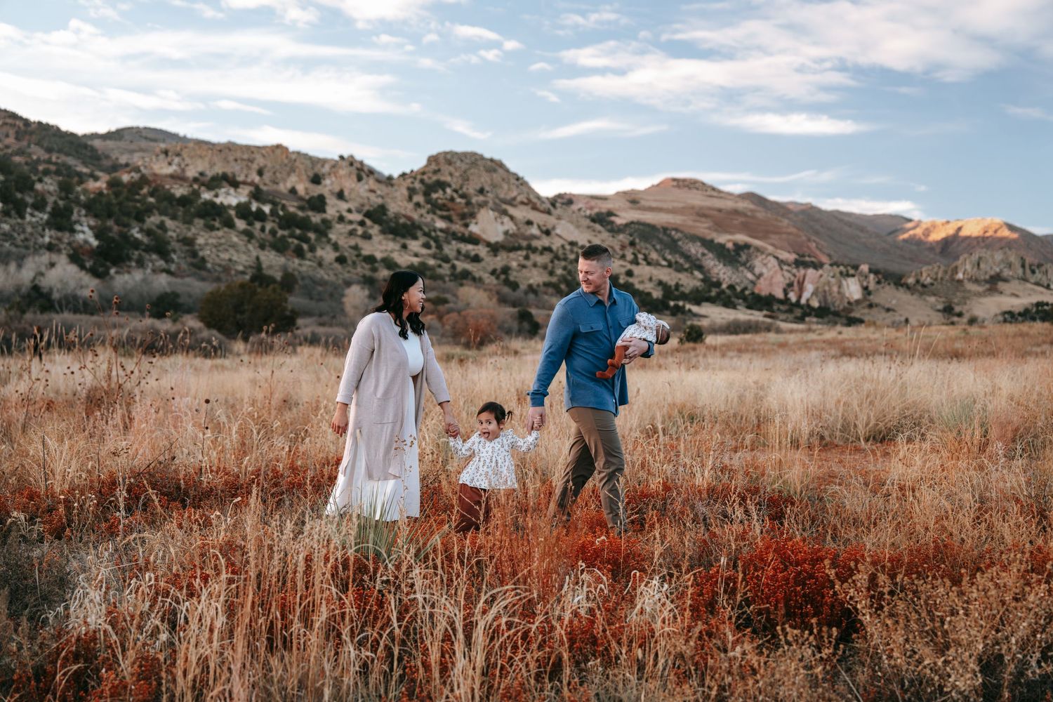 Young family of four walking through golden autumn field with rocky mountain landscape in background.