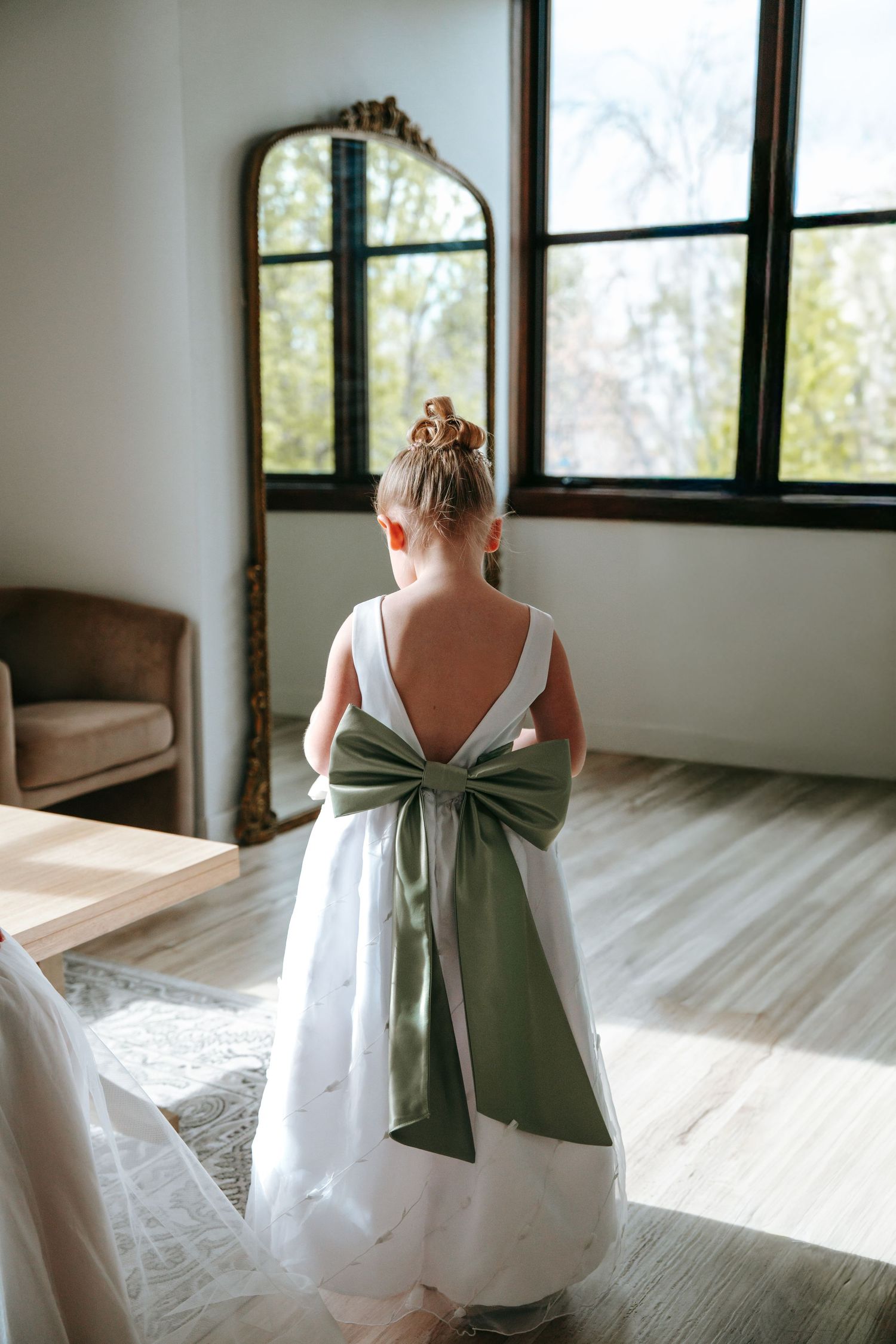 Flower girl in white dress with sage green bow standing near gold mirror in bridal suite.