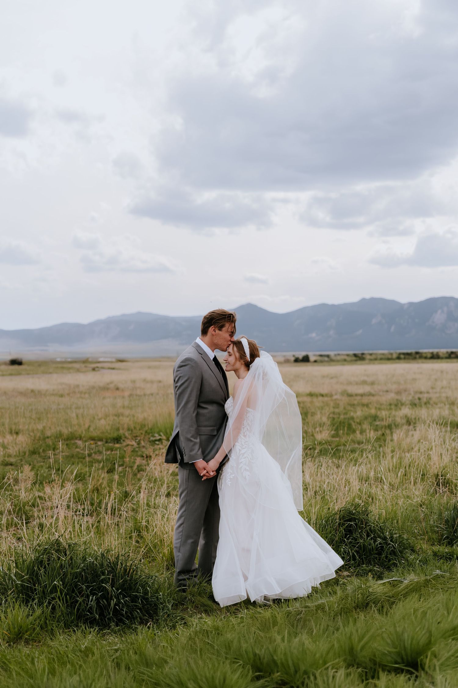 Bride and groom share a kiss in a golden meadow with mountain range and dramatic cloudy sky behind them.