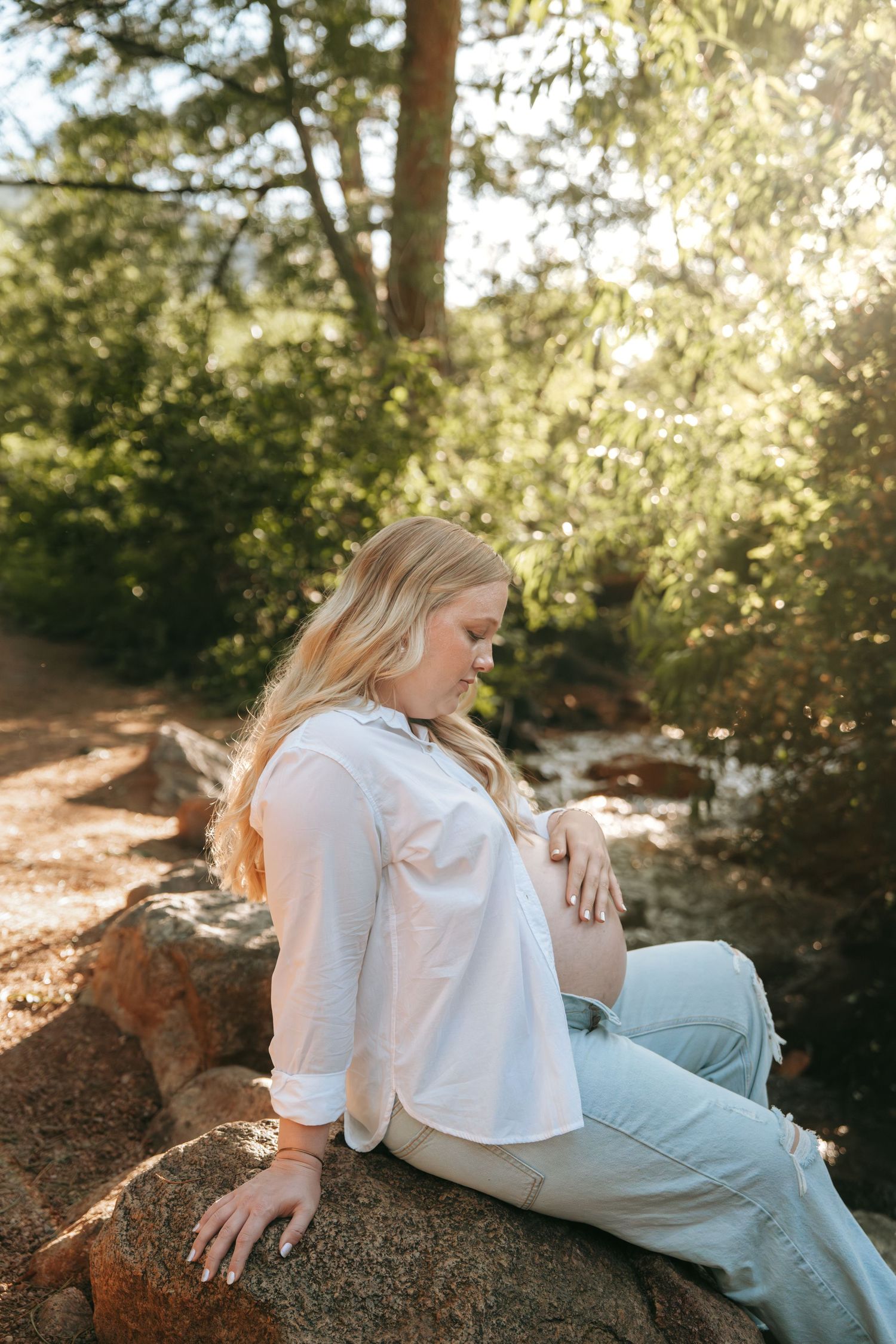 Pregnant blonde woman in white shirt sits on rocks in a sunny forest setting.