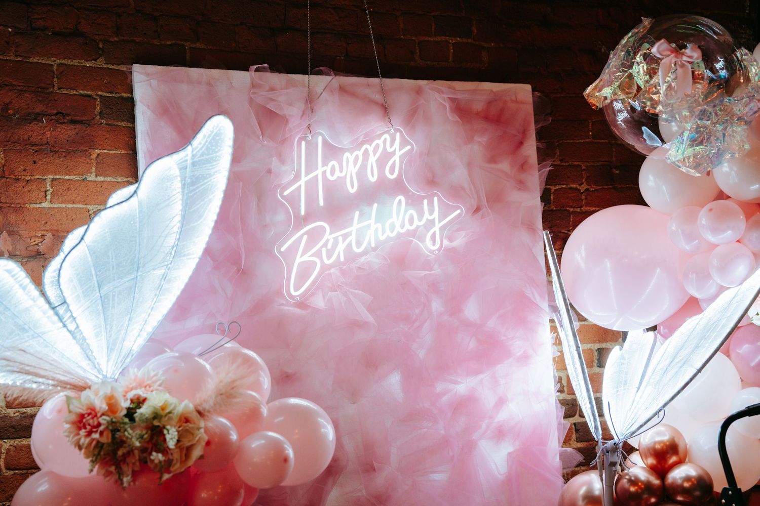 Pink neon Happy Birthday sign with butterfly lights and balloon decorations against a brick wall.