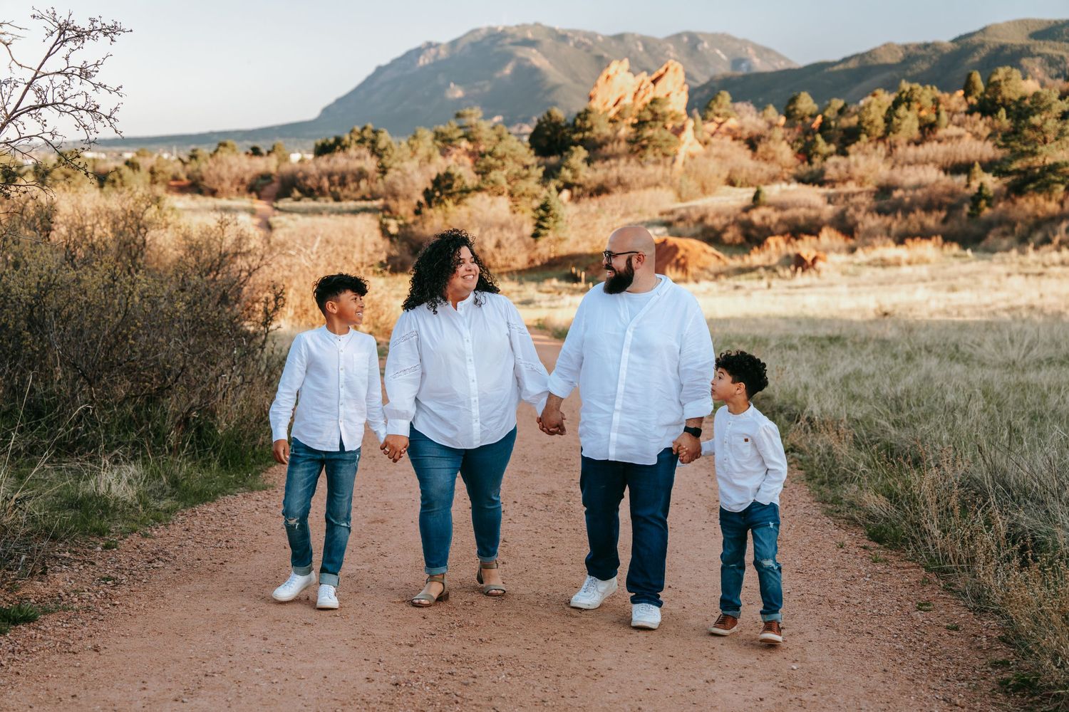 Happy family of four in matching white shirts walking hand in hand on a dirt path near rocky mountains.