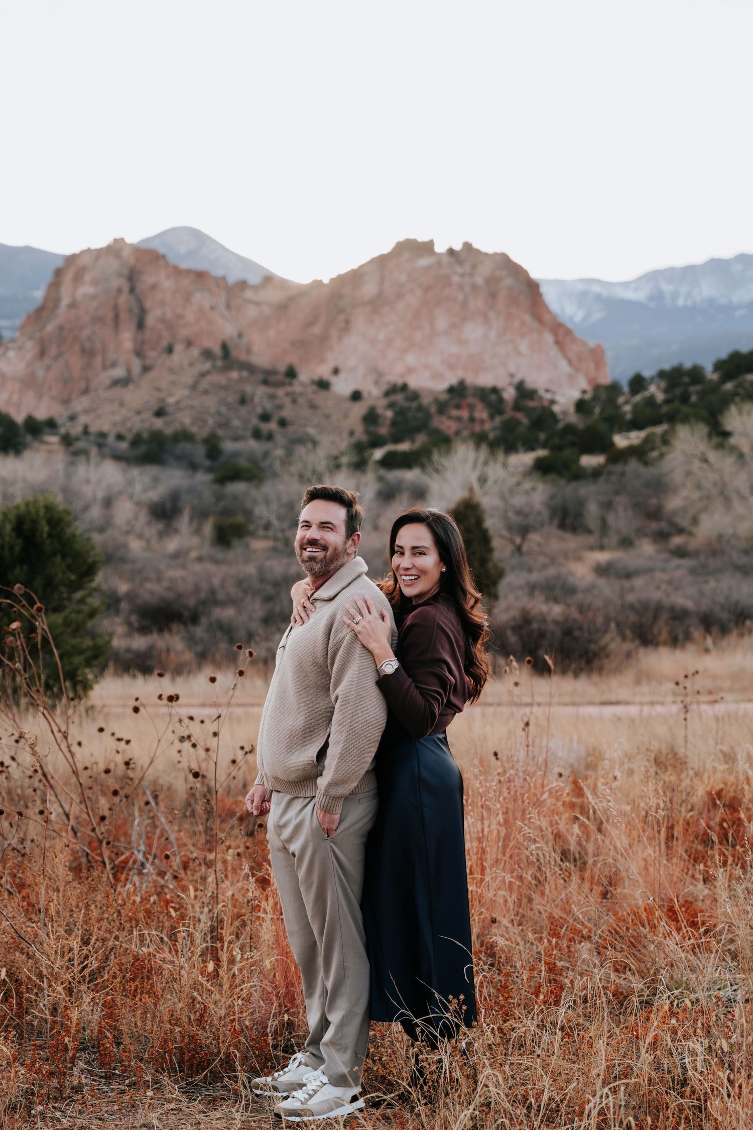 Smiling couple embracing in a dry field with dramatic red rock mountain formations in background.